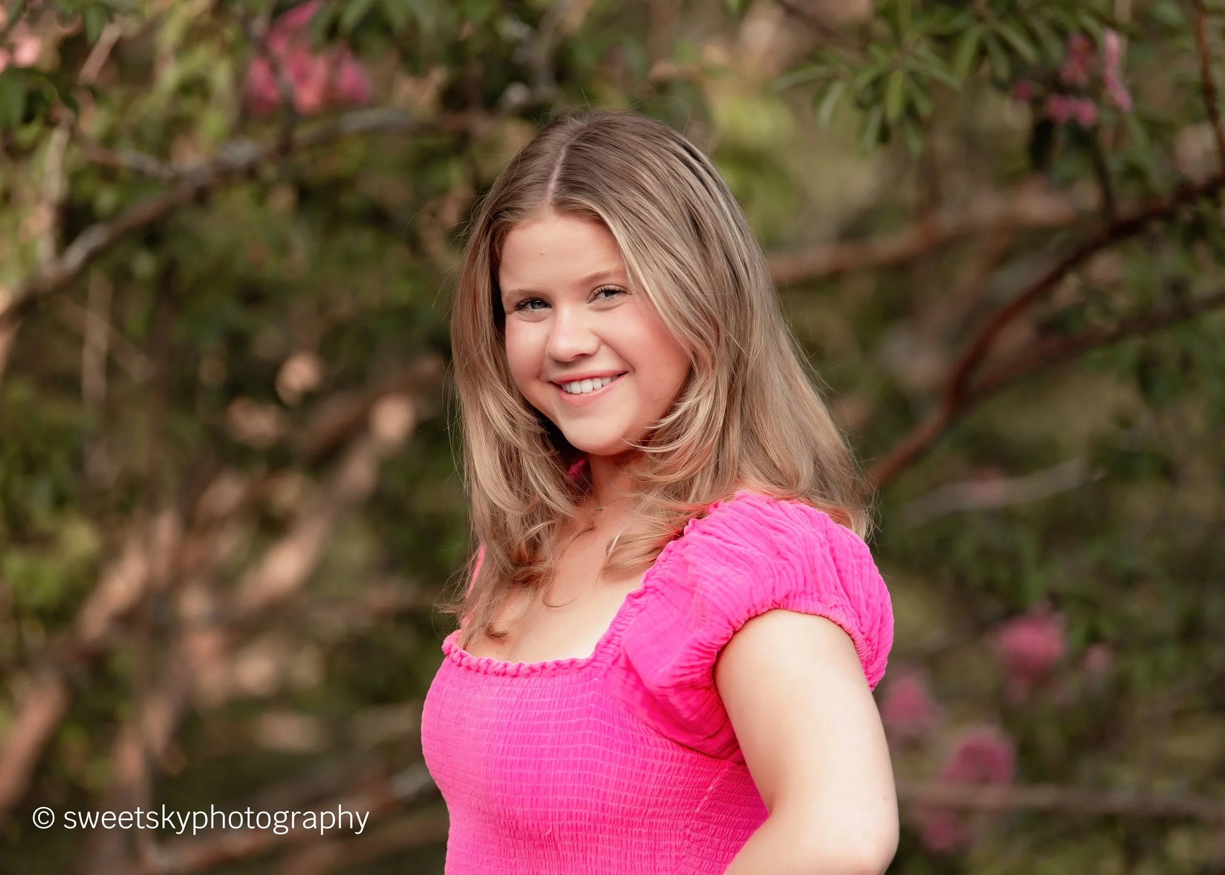 A young woman with blonde hair smiling outdoors, wearing a pink short-sleeve top, with a background of green foliage and pink flowers.
