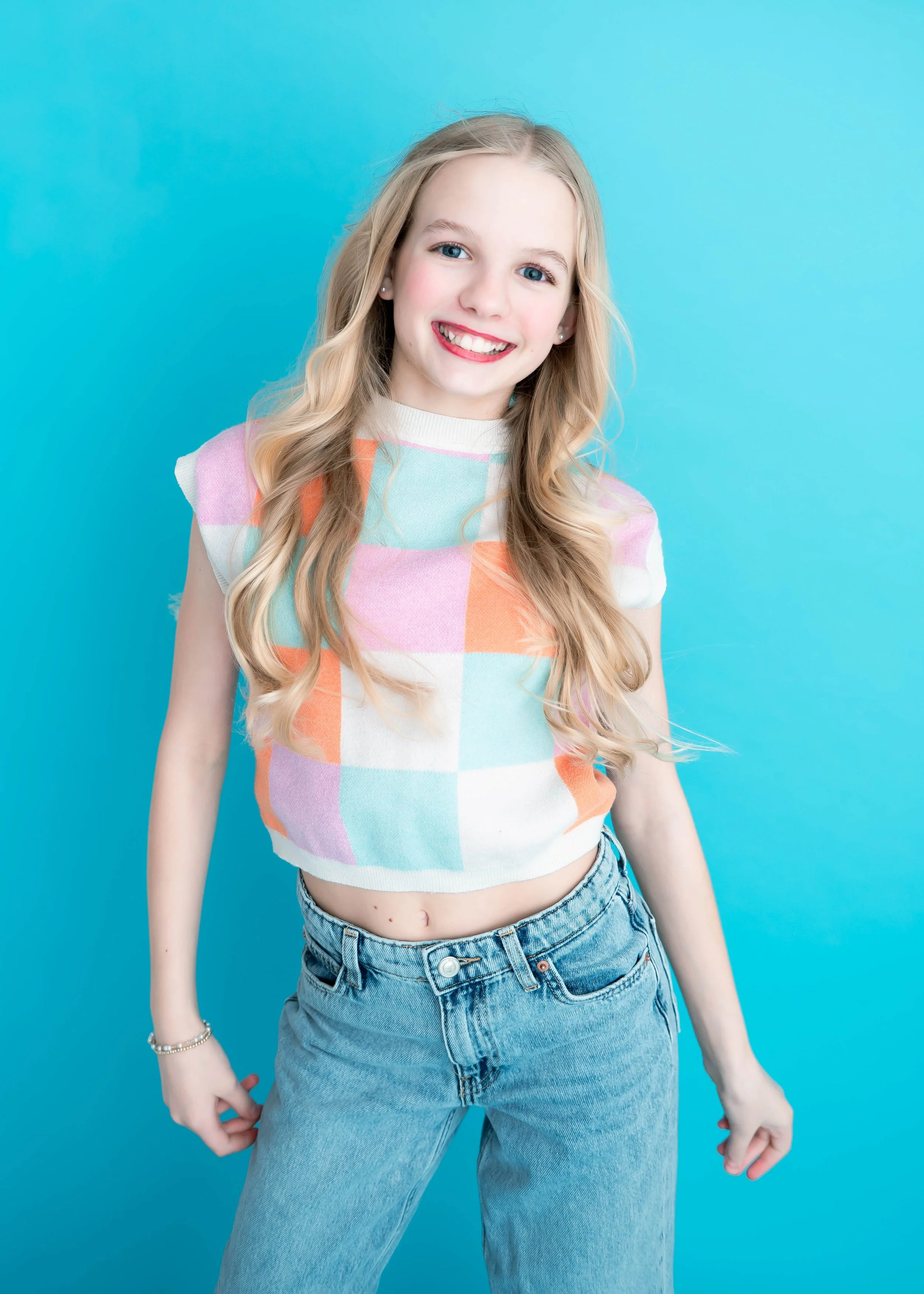 A young girl with long blonde hair and blue eyes smiling, wearing a pastel checkered sleeveless top and jeans, standing against a light blue background.