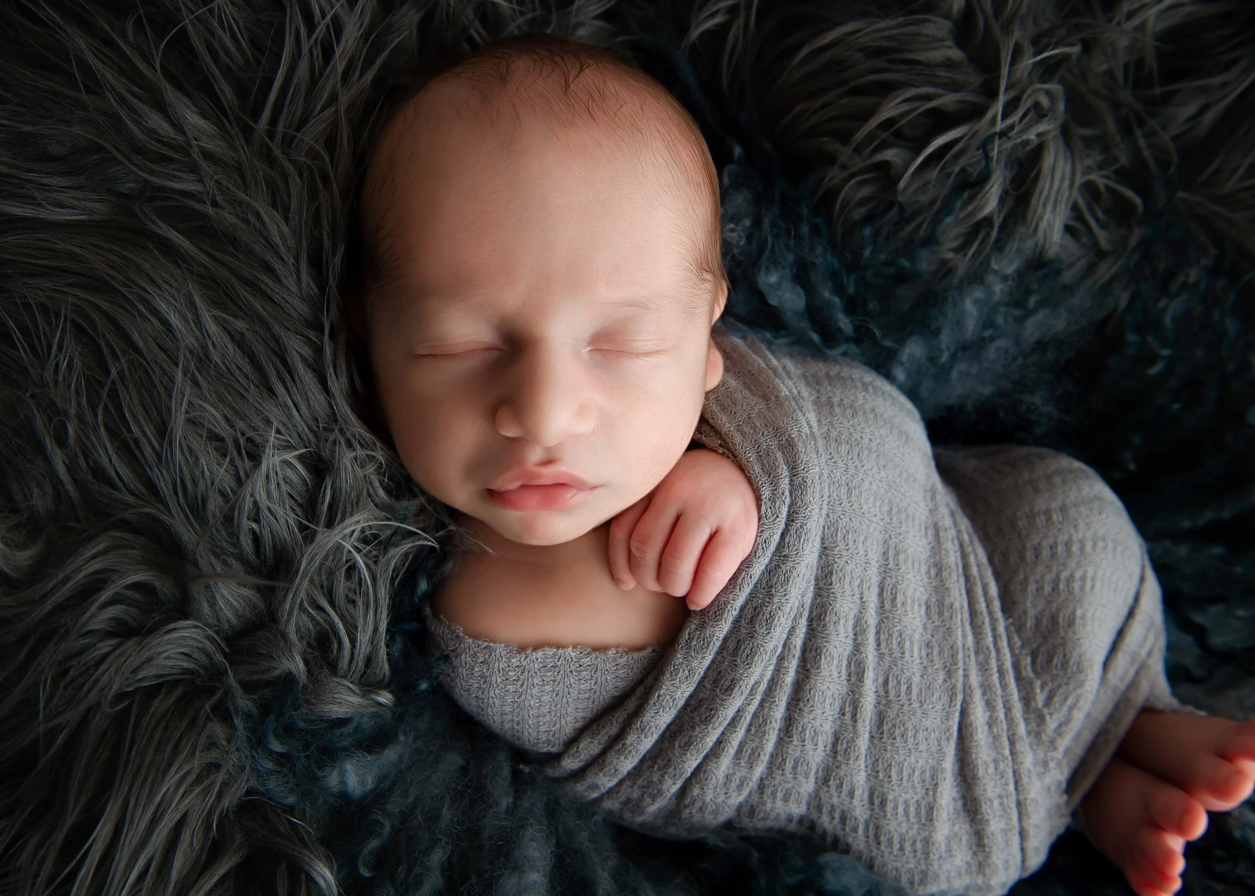 Close-up of a sleeping newborn baby with a serene expression, resting on a furry dark gray blanket, wearing a knitted gray outfit.