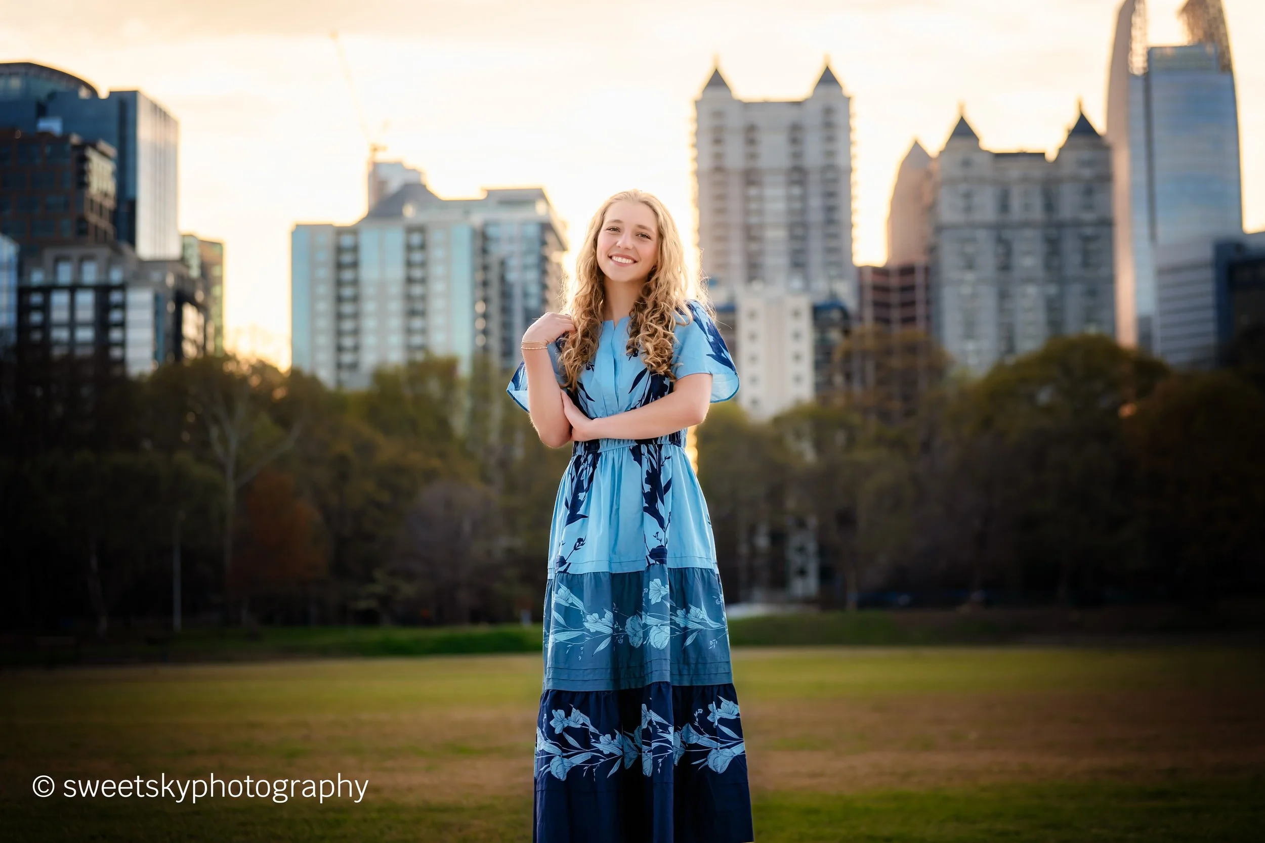 A smiling woman in a blue floral dress standing in a park with city buildings in the background at sunset.
