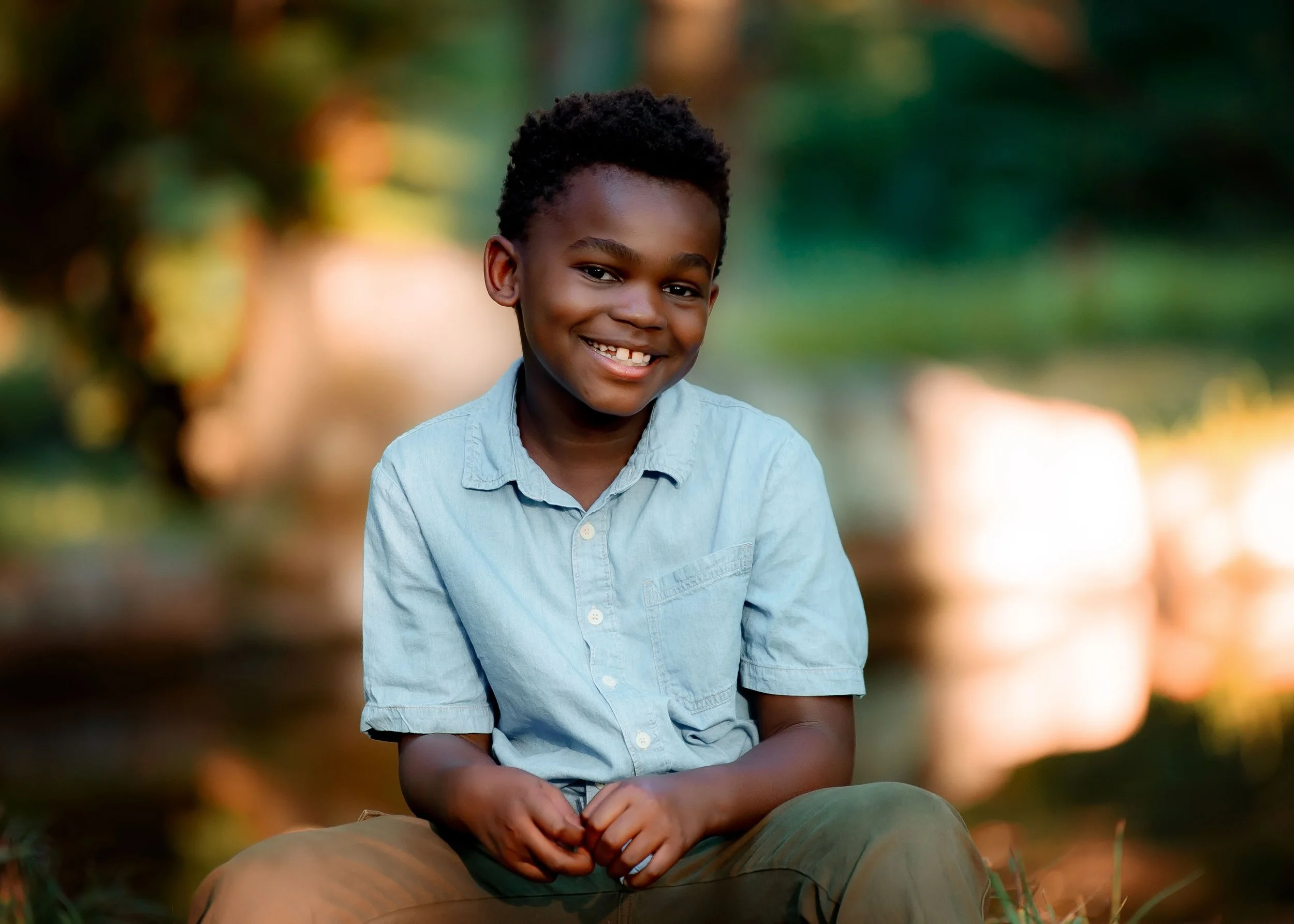 A smiling young boy with dark hair sitting outdoors by a body of water during sunset.