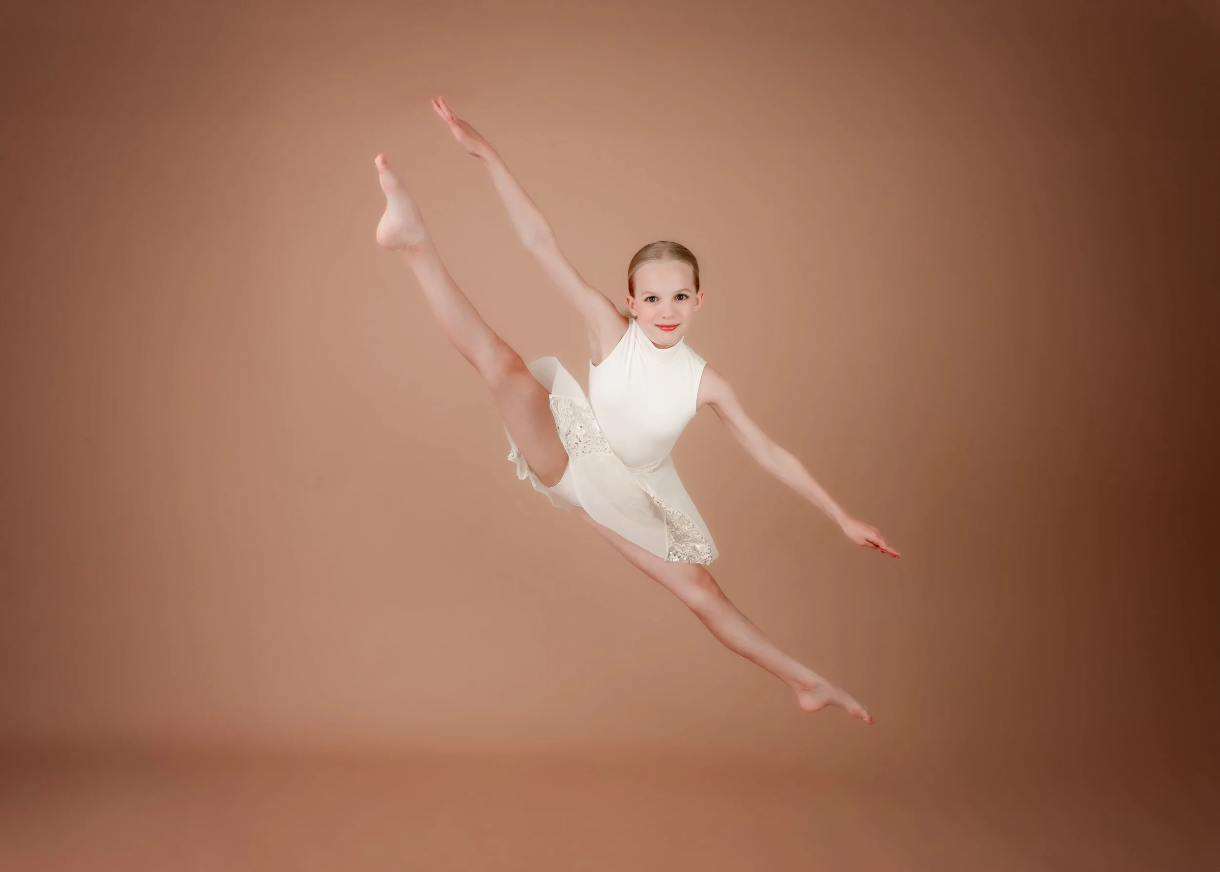 Young girl in a white dance costume performing a split leap with arms extended, against a brown background.