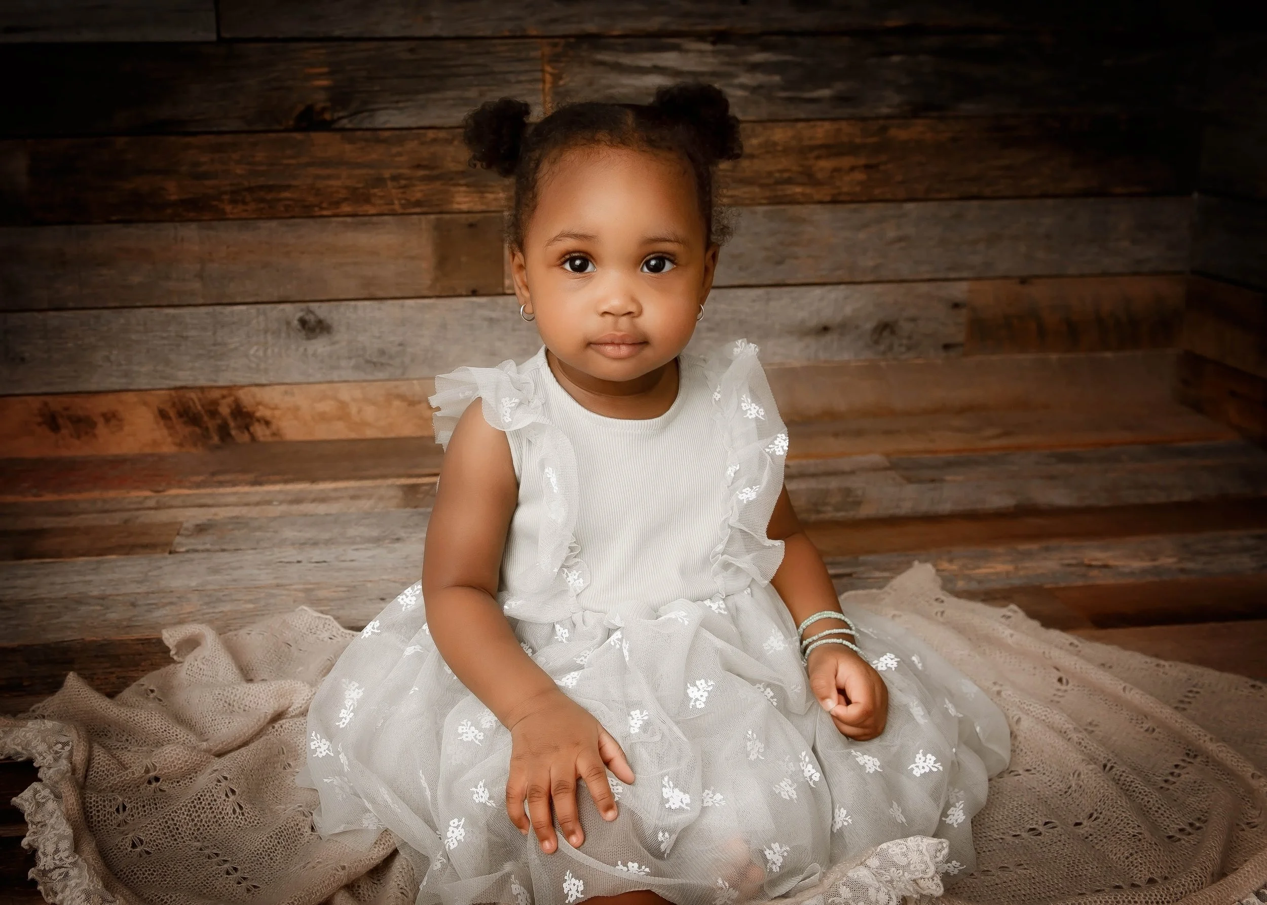 A young girl with dark hair styled in two puffs, wearing a white dress with lace details, sitting on a lace blanket in front of a wooden backdrop.