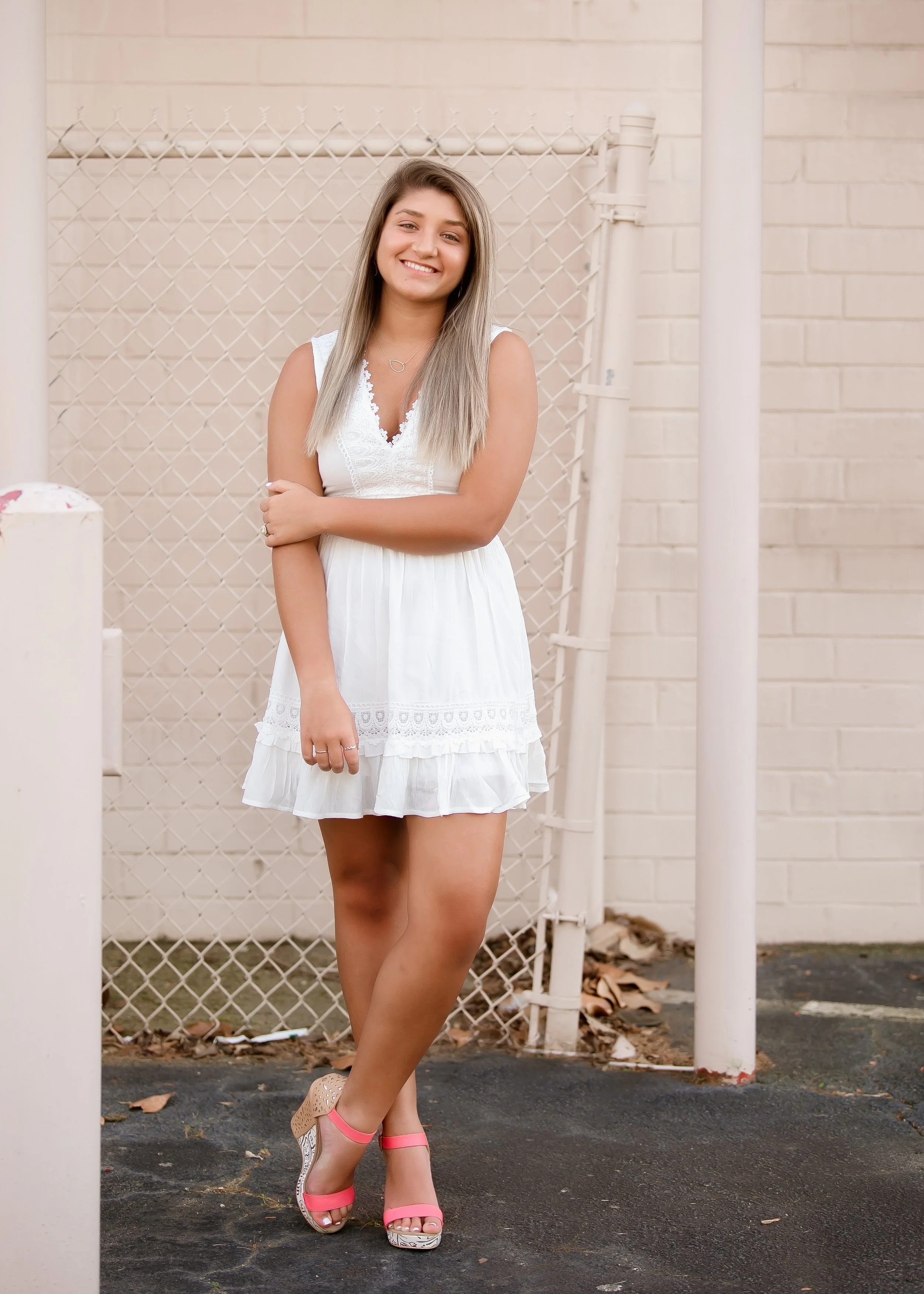 A young woman with long hair smiling and standing outdoors near a beige brick wall and chain-link fence, wearing a white sleeveless dress and pink high-heeled sandals.