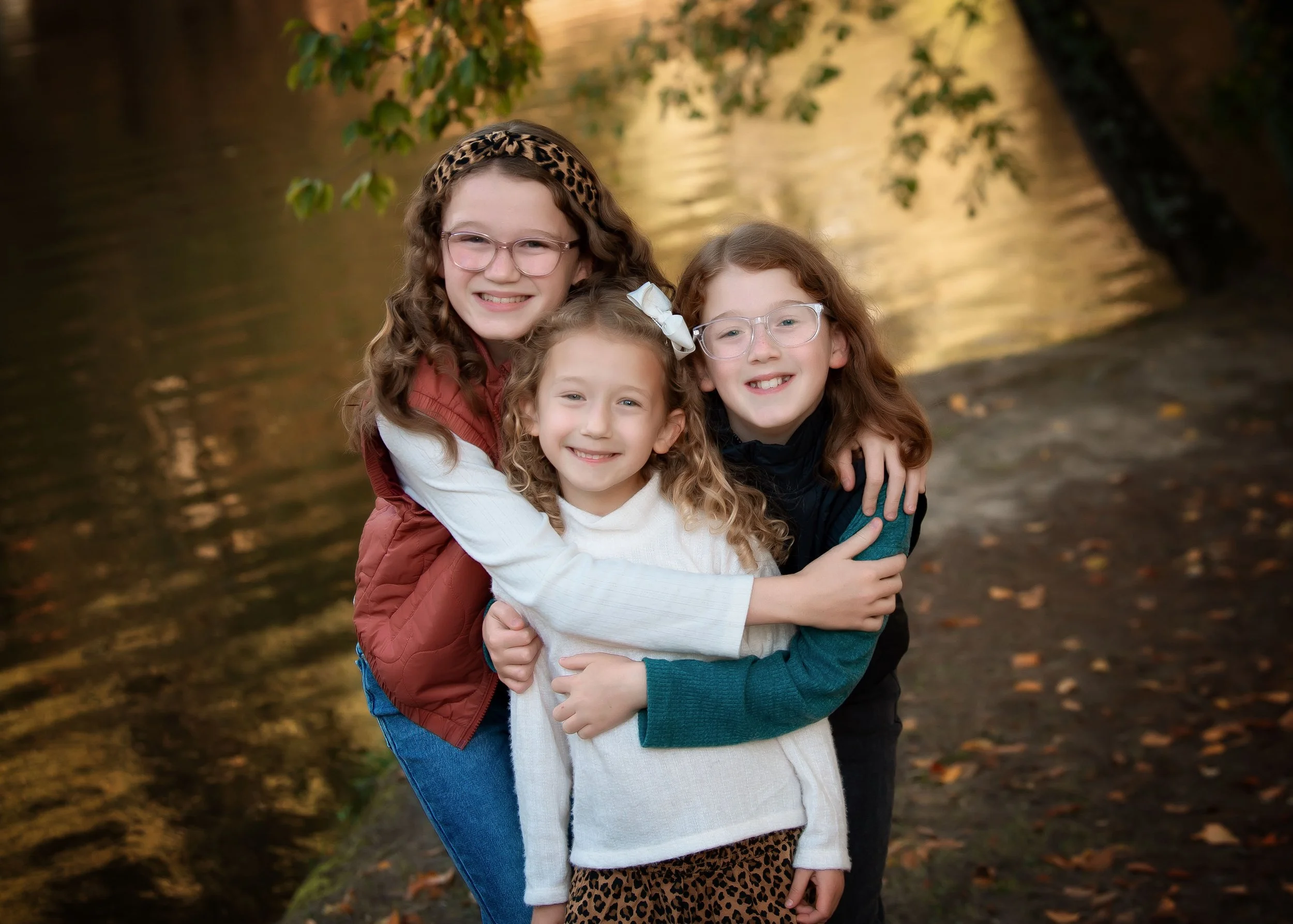 Three girls hugging outdoors near a river at sunset, with trees and leaves on the ground.