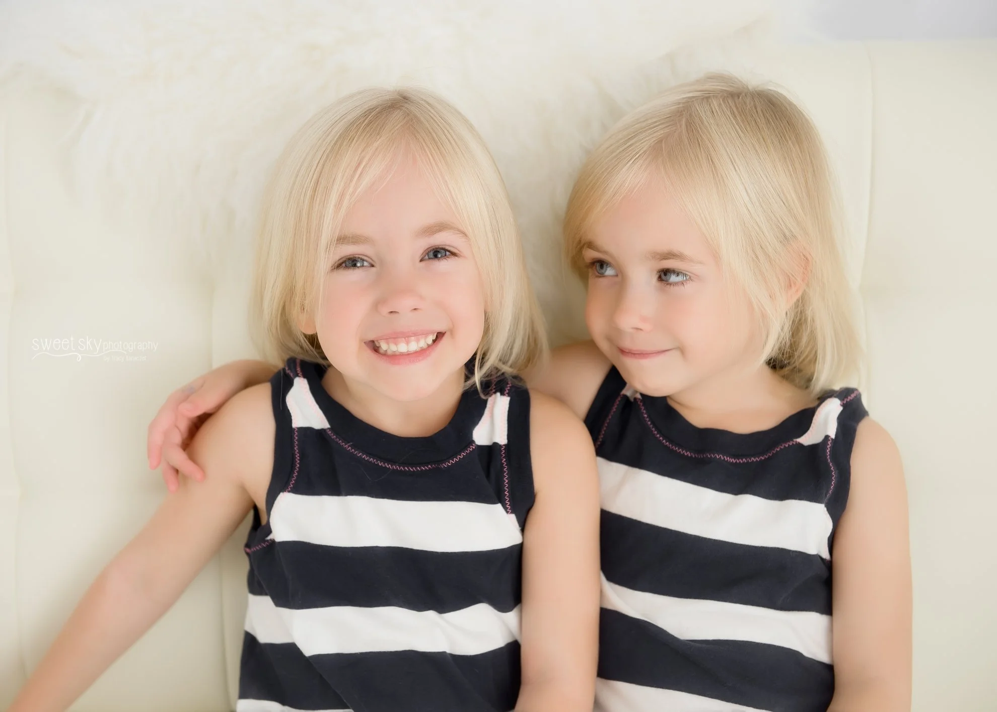 Two young blonde-haired girls sitting on a cream-colored couch, wearing matching black and white striped sleeveless dresses, smiling and relaxing together.