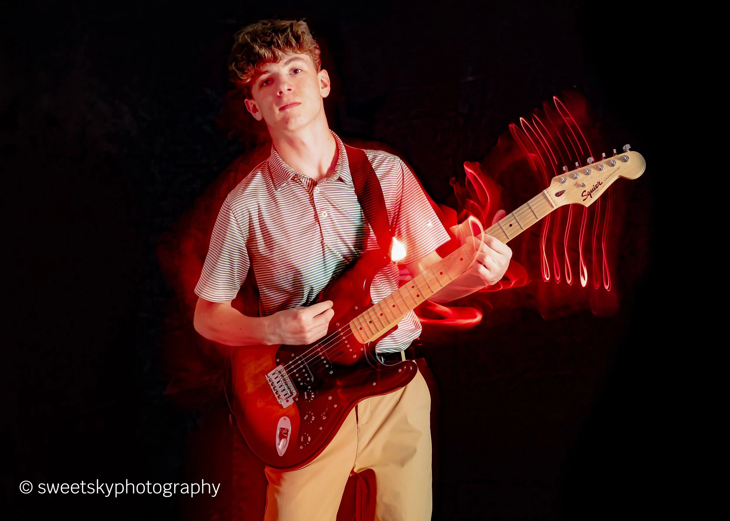 Young man with curly hair wearing a striped shirt and beige pants playing an electric guitar against a black background with red light trails.