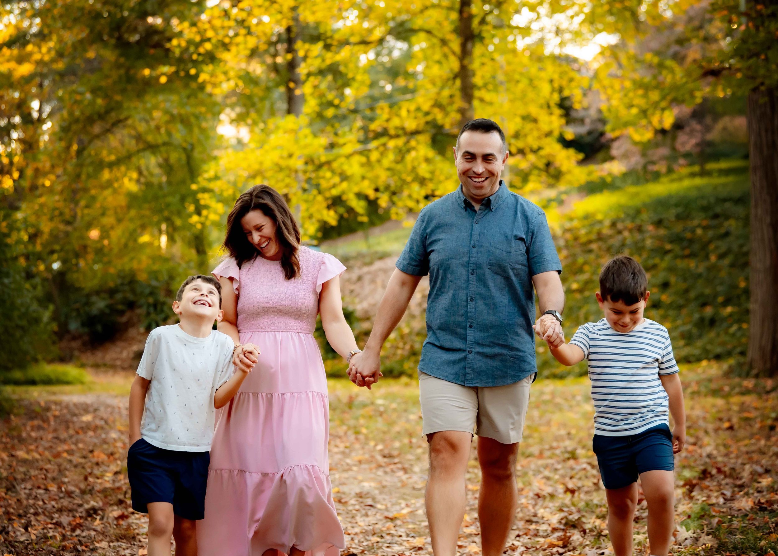 A family of four walking hand in hand through a park during autumn, surrounded by yellow and green leaves on trees.