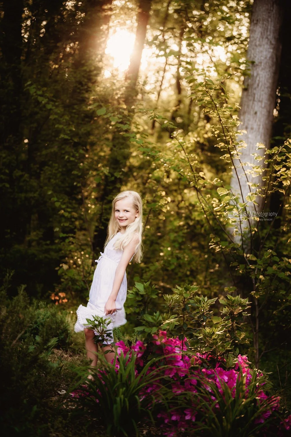 A young girl with long blonde hair in a white dress standing outdoors in a wooded area with sun shining through the trees and pink flowers in the foreground.