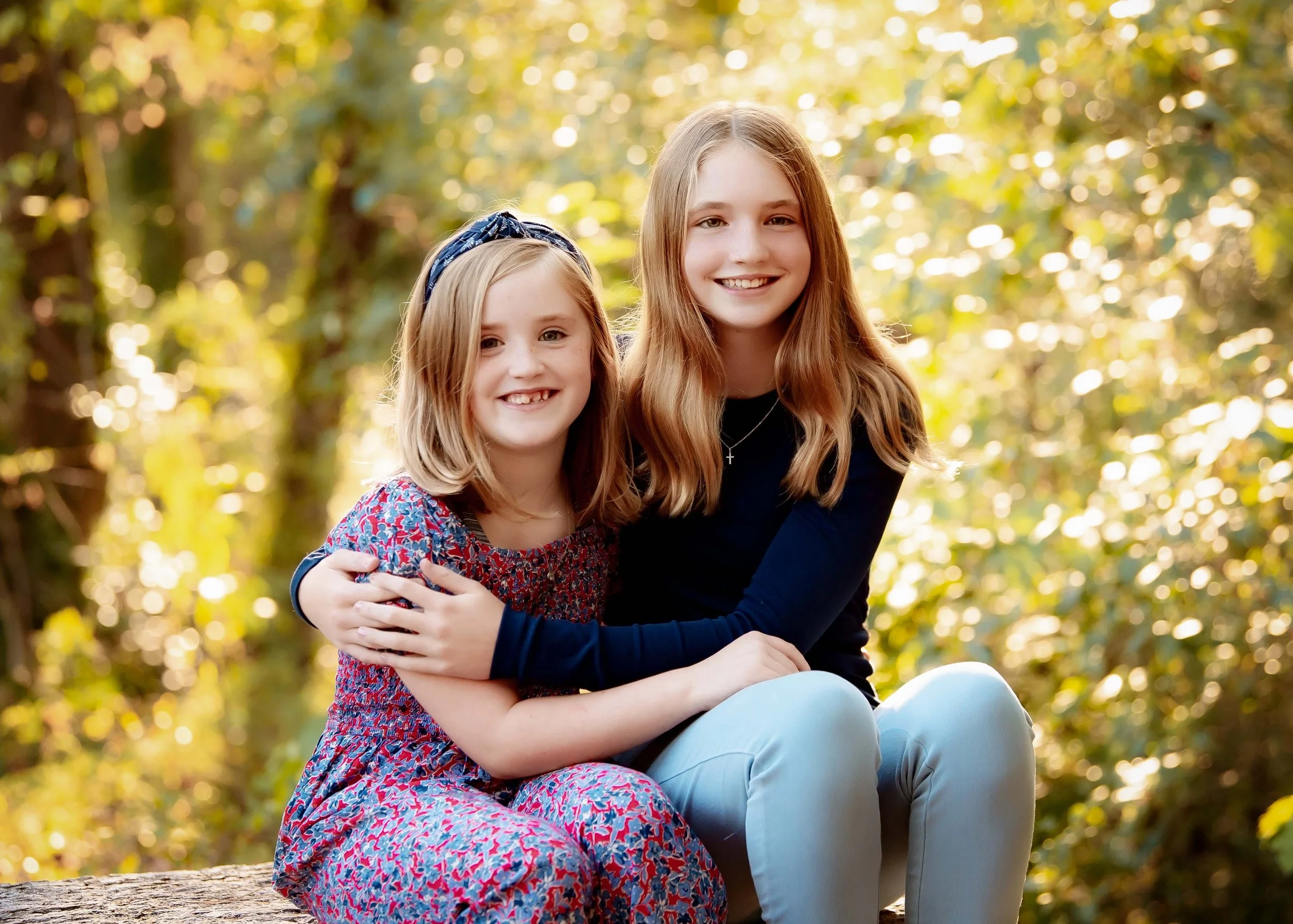 Two young girls sitting outdoors on a log with a blurred forest background, smiling at the camera. One girl is wearing a floral dress and headband, the other a black top and light-colored pants.