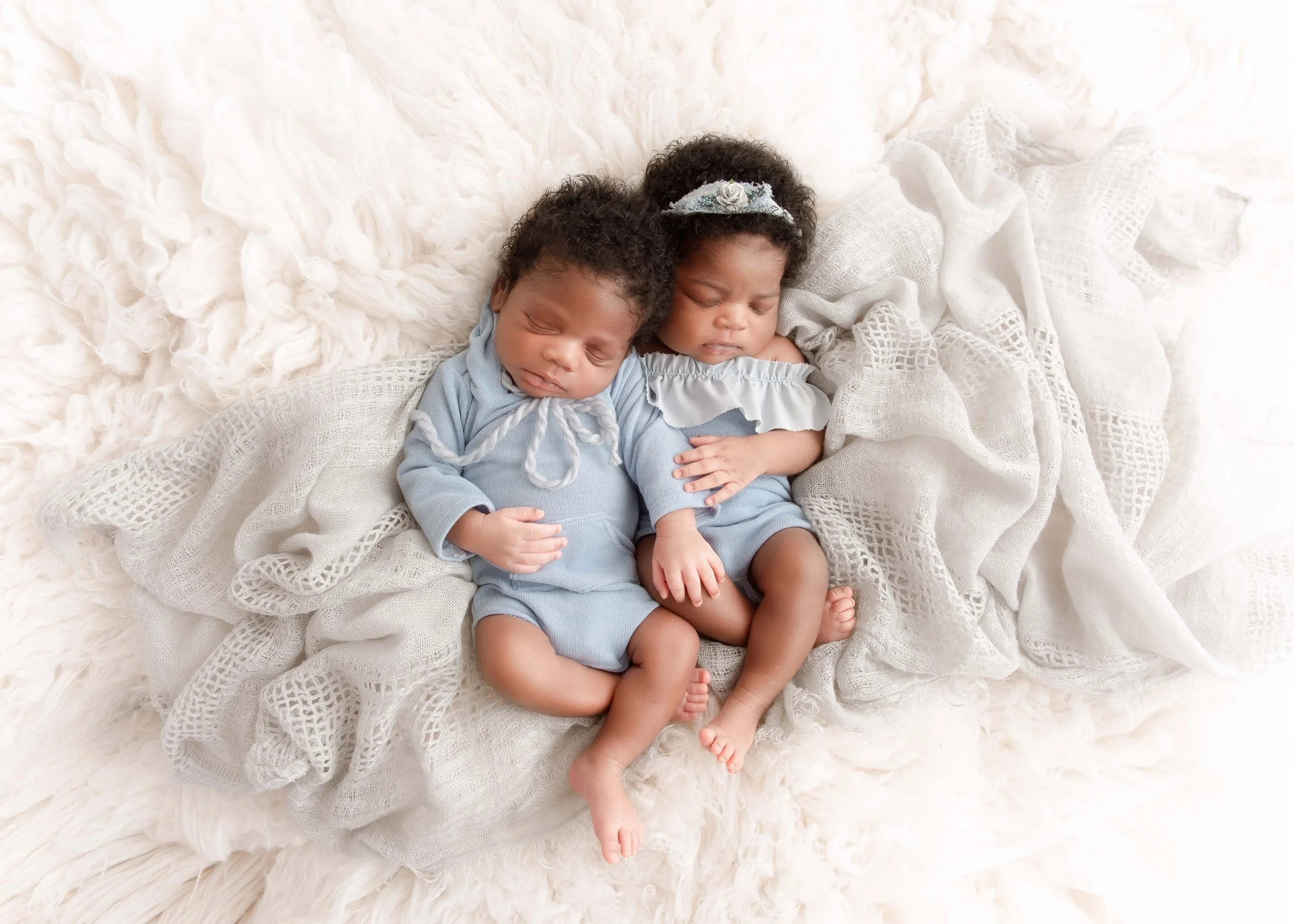 Two sleeping babies, a boy and a girl, cuddled together on a soft, textured blanket with a light-colored, lacy fabric surrounding them.