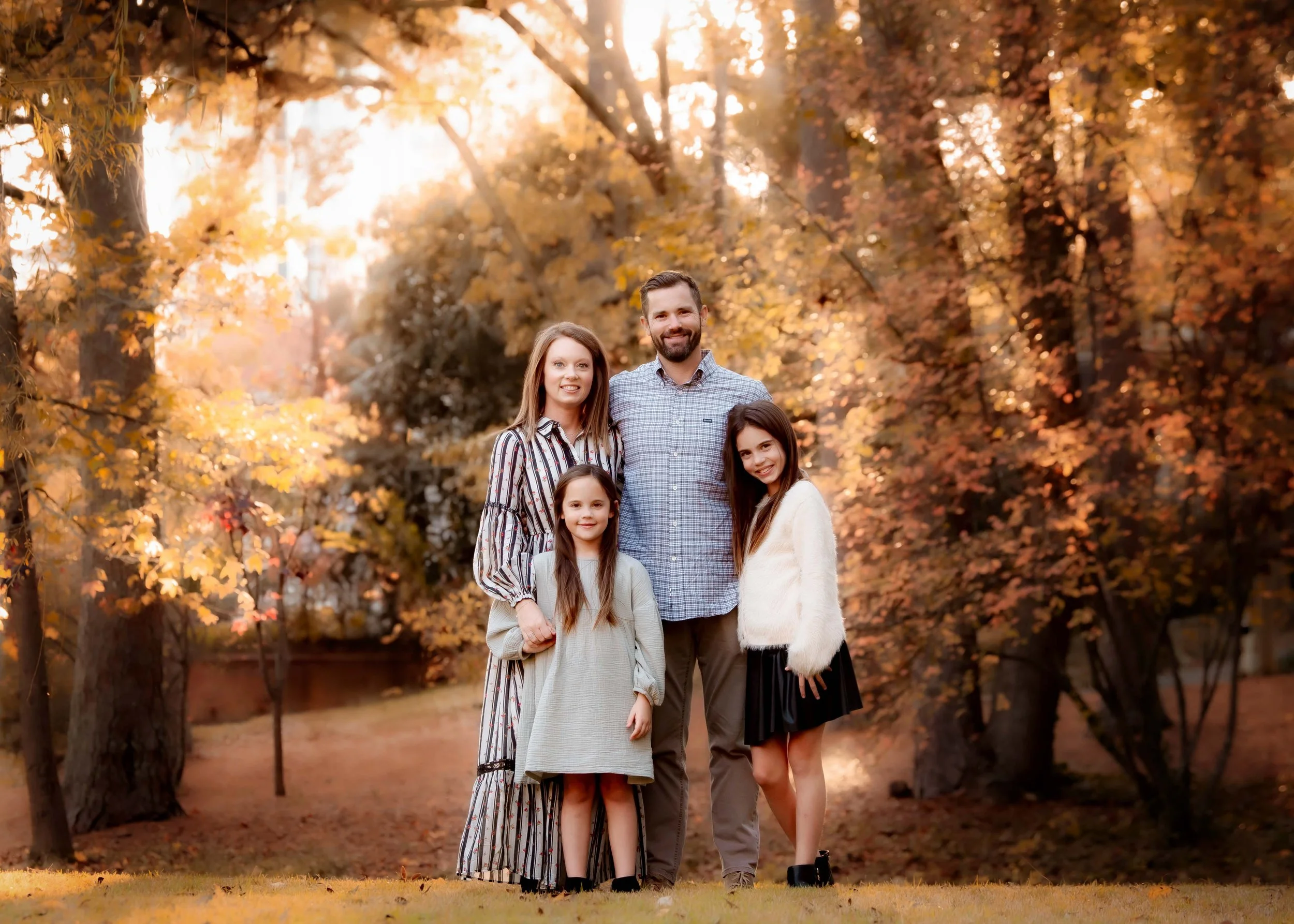 A family of five posing outdoors in an autumn park with trees and fallen leaves, sunlight shining through the leaves.