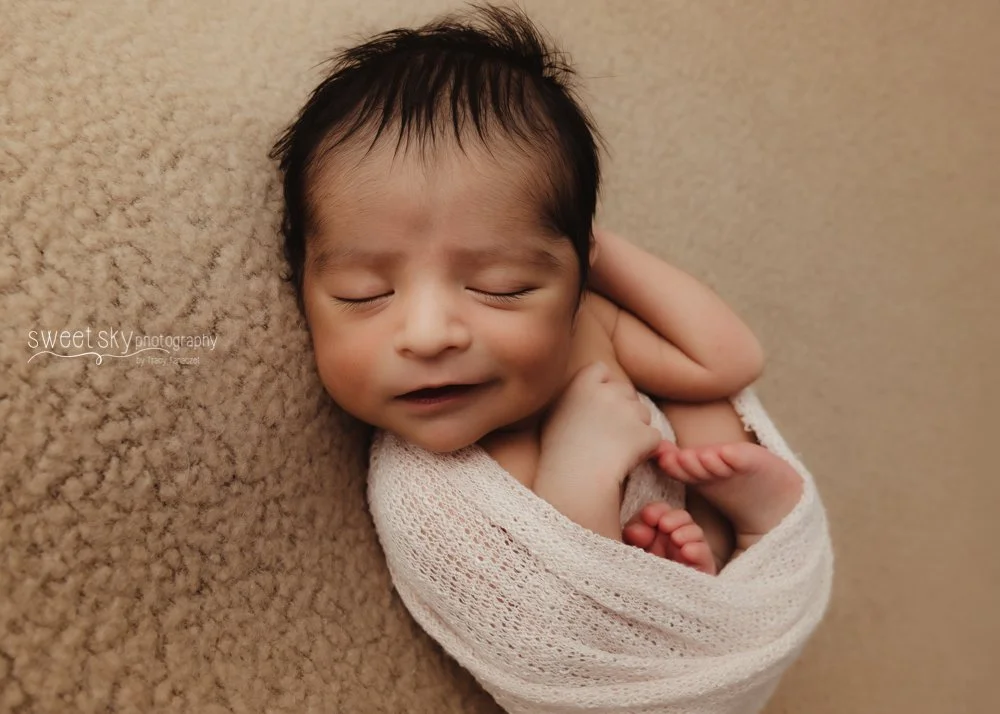 A newborn baby with dark hair sleeping on a beige blanket, wrapped in a white blanket, with a peaceful expression.