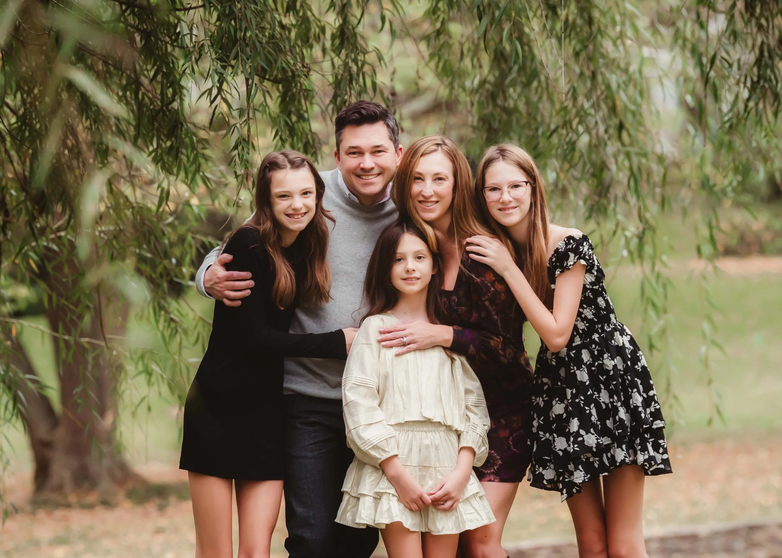 A family of six posing outdoors among trees with green leaves, smiling and hugging each other.