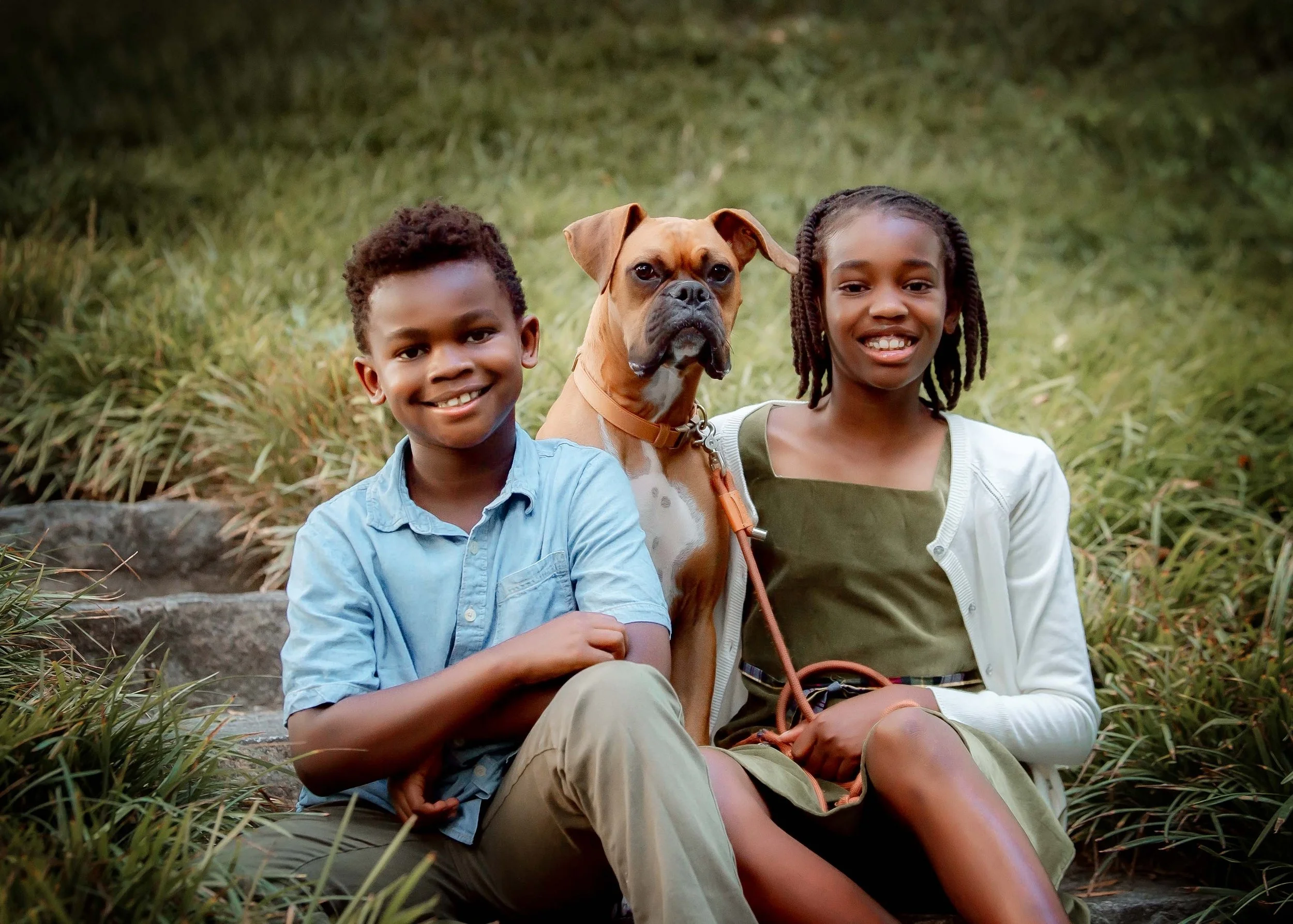 A boy, a girl, and a brown dog sitting on steps outdoors, surrounded by grass, smiling at the camera.
