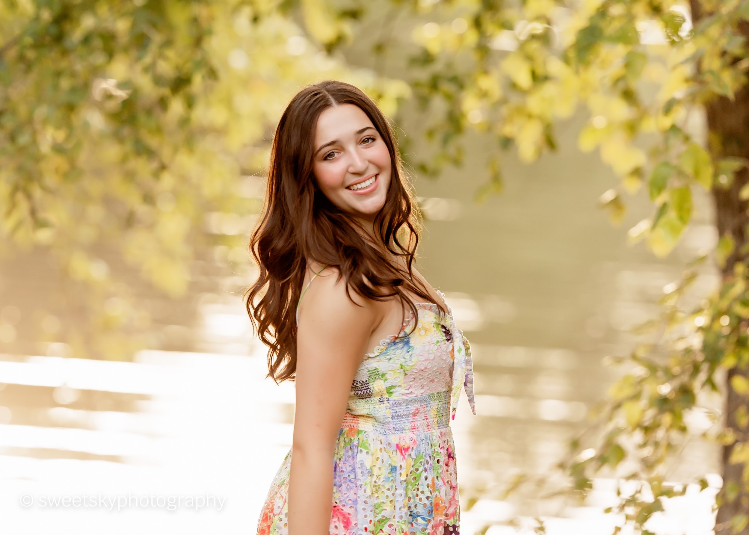 A young woman with long brown hair, wearing a colorful, patterned summer dress, smiling and standing outdoors near a body of water with trees in the background.