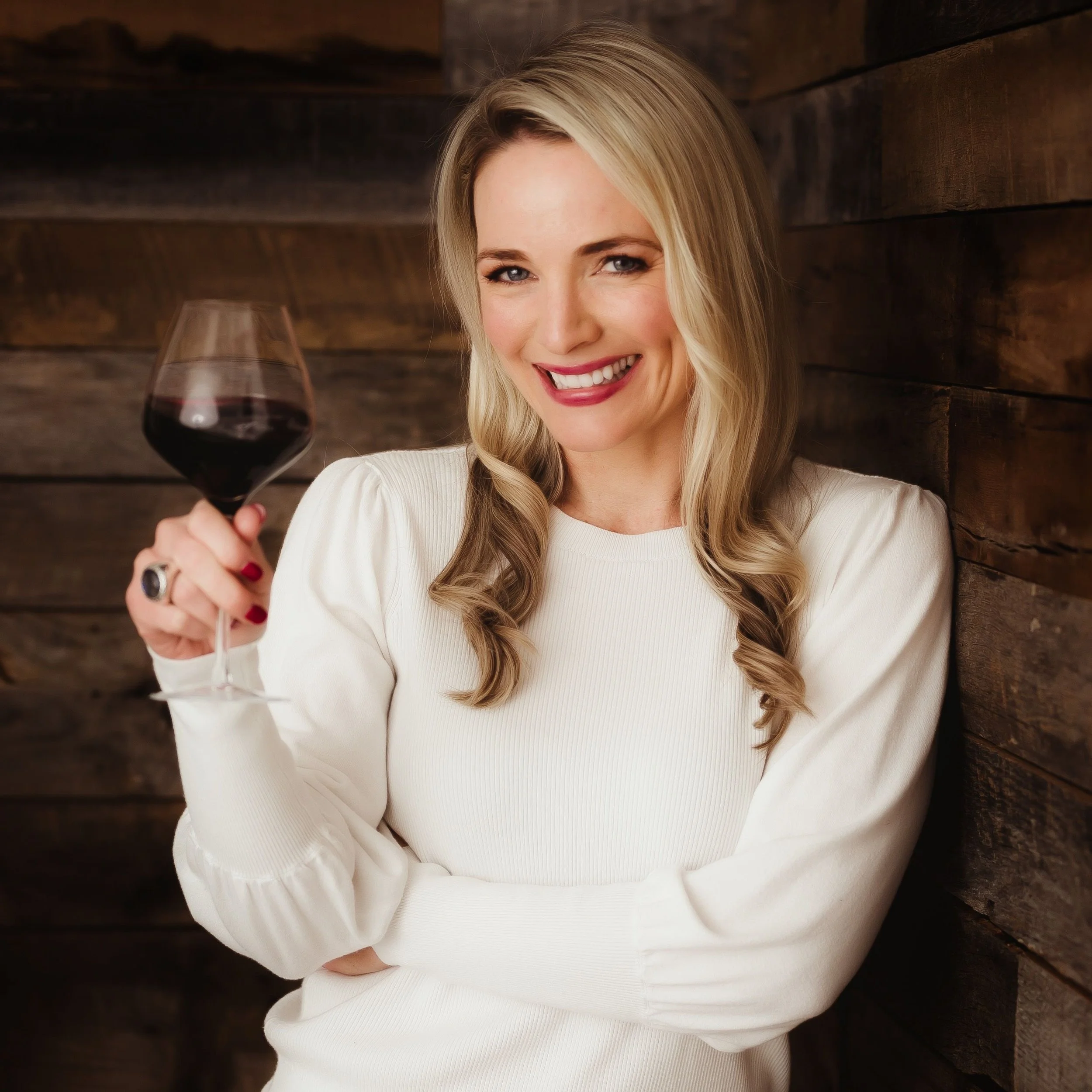 A woman holding a glass of red wine, smiling, and wearing a white top, standing against a wooden wall.