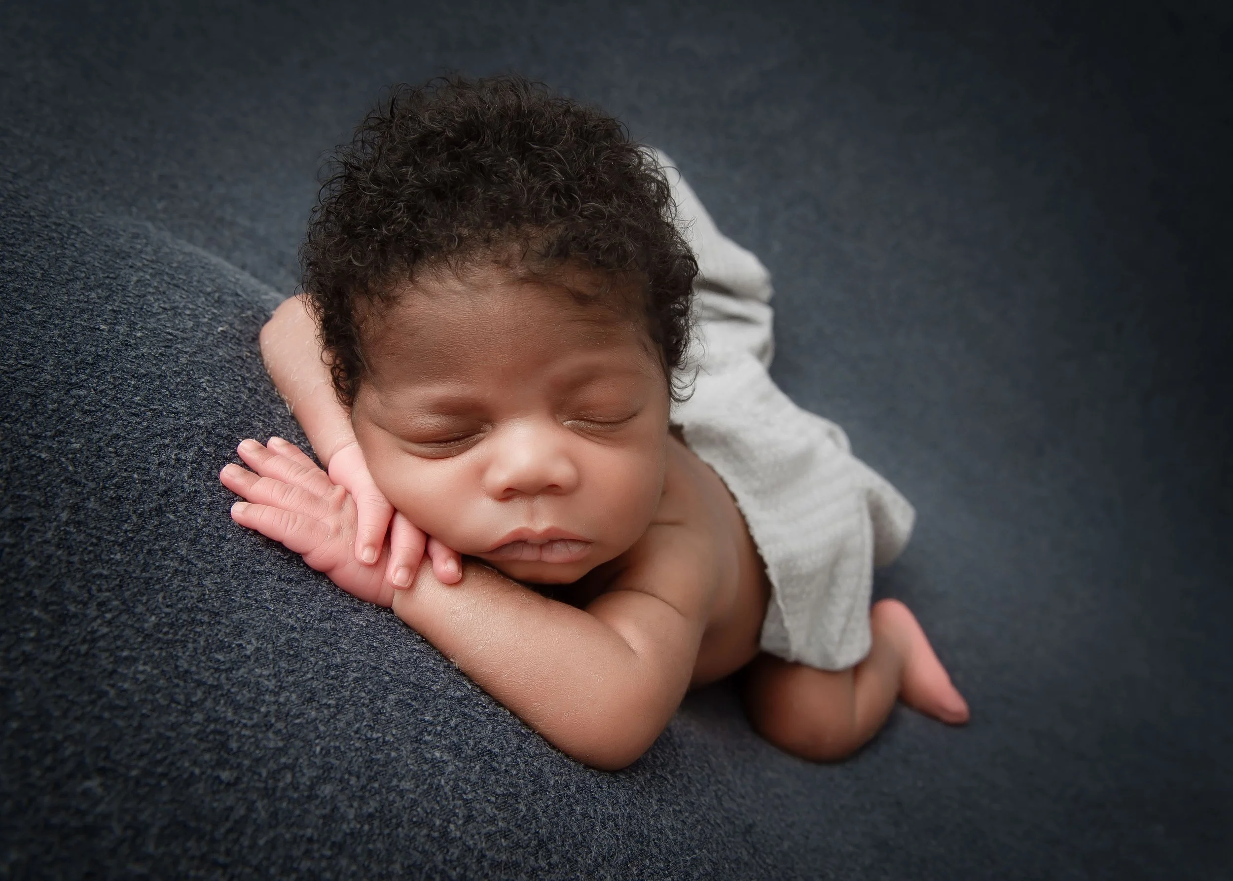 A sleeping baby with curly hair resting on a dark gray surface, with one hand under their chin and the other on their cheek.