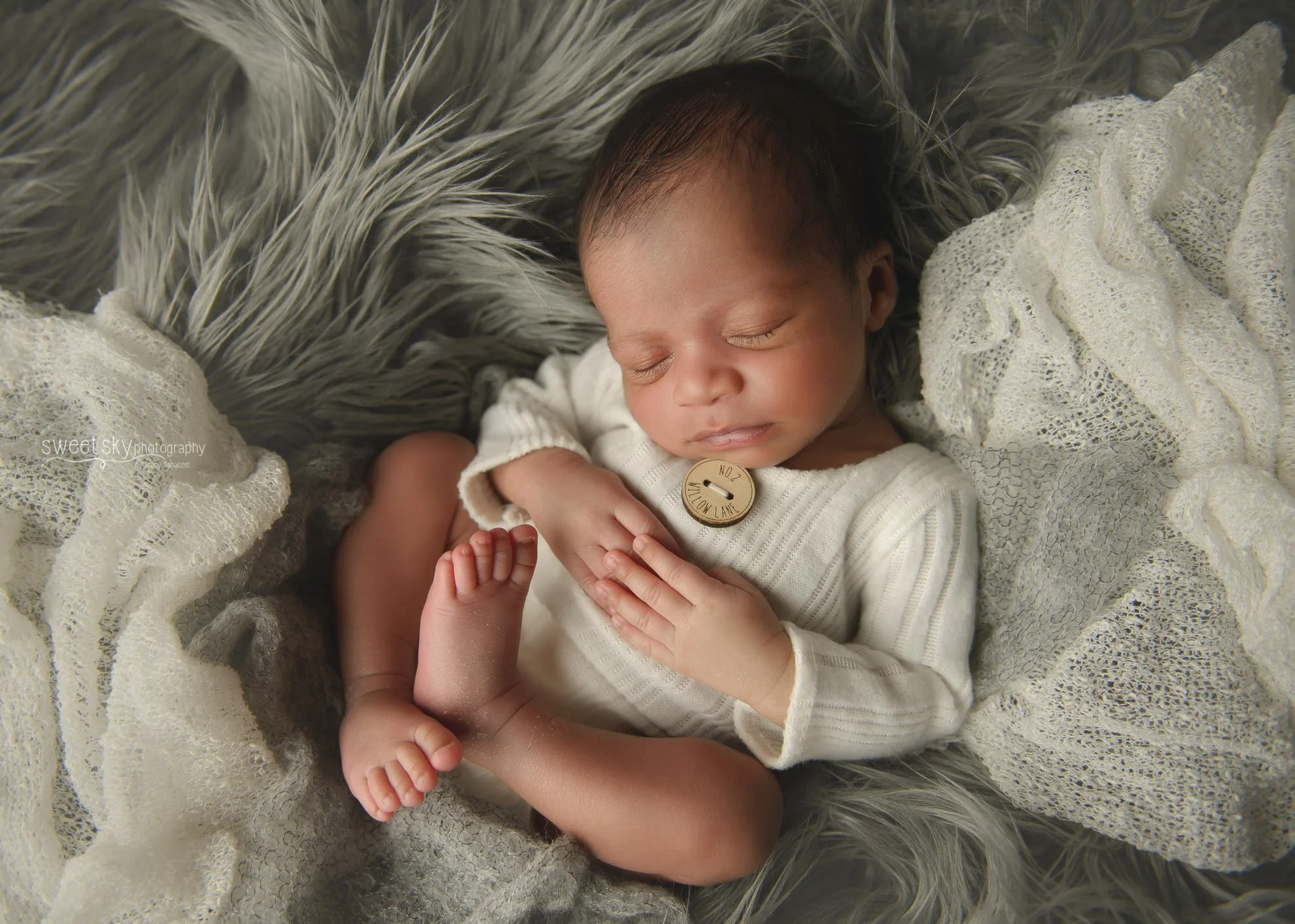 Sleeping baby with curly hair, dressed in white, laying on gray faux fur and lace fabrics, with a wooden button near neck.
