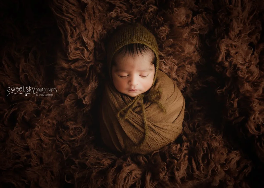 A newborn baby peacefully sleeping, wrapped in a brown blanket and wearing a matching hat, lying on a textured brown fur blanket.