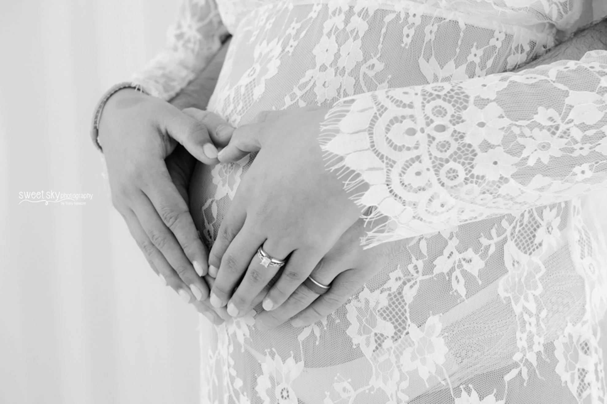 Close-up of a woman’s hands forming a heart shape over her pregnant belly, wearing a lace dress and wedding rings.