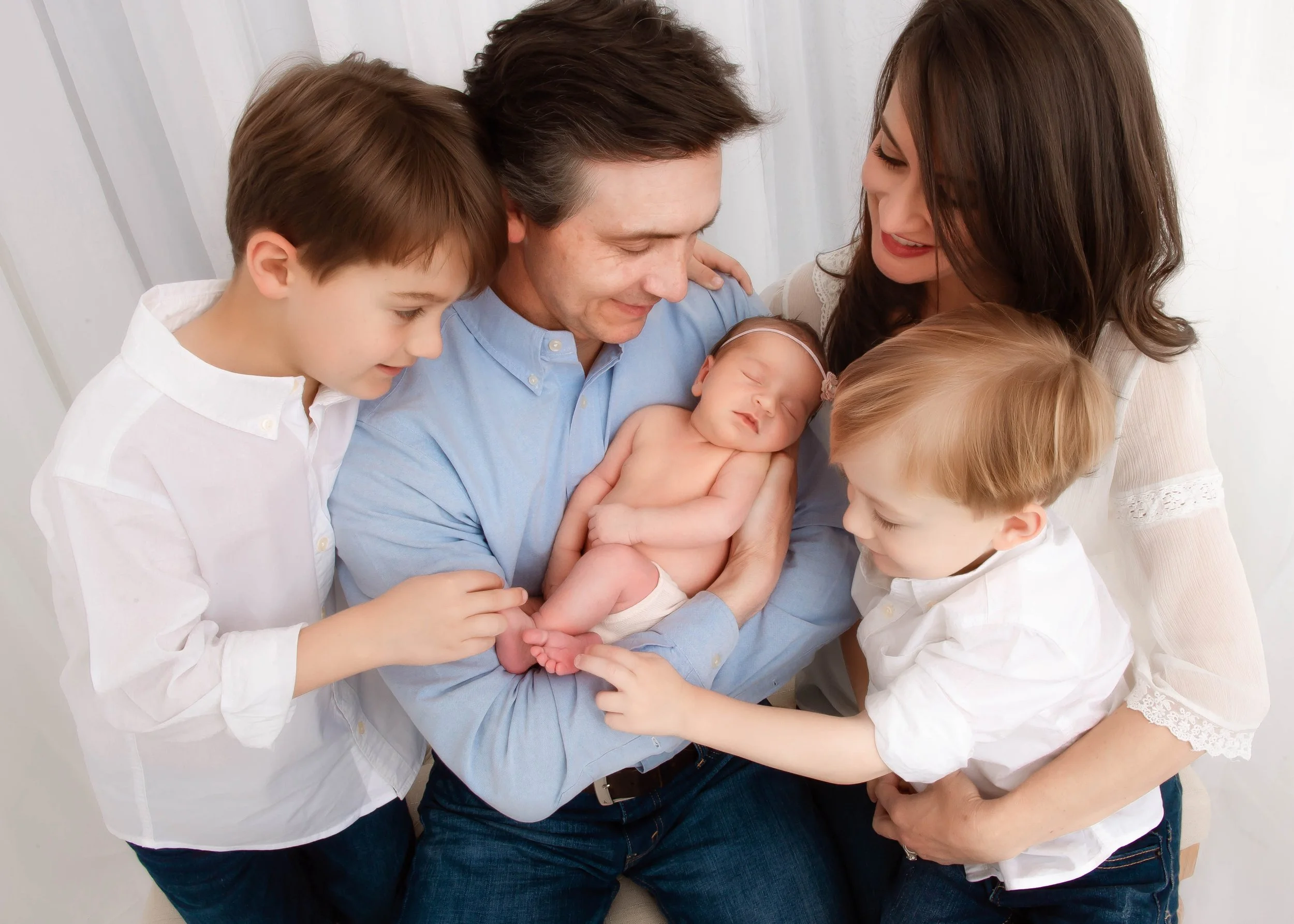 Family holding newborn baby, surrounded by two young boys and a woman, in a bright white room