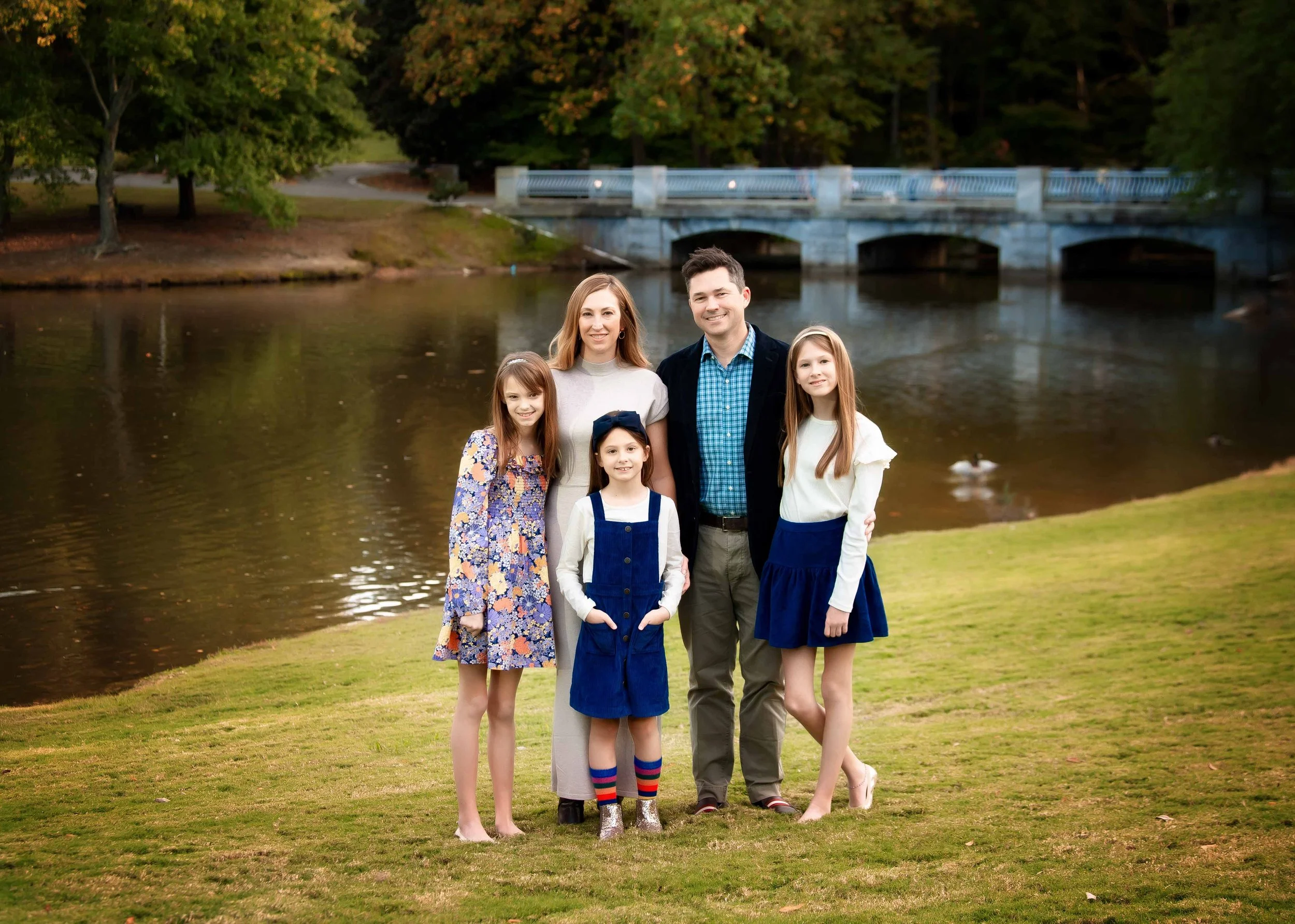 Family photo of two adults and three young girls standing on grass near a pond, with trees and a bridge in the background.
