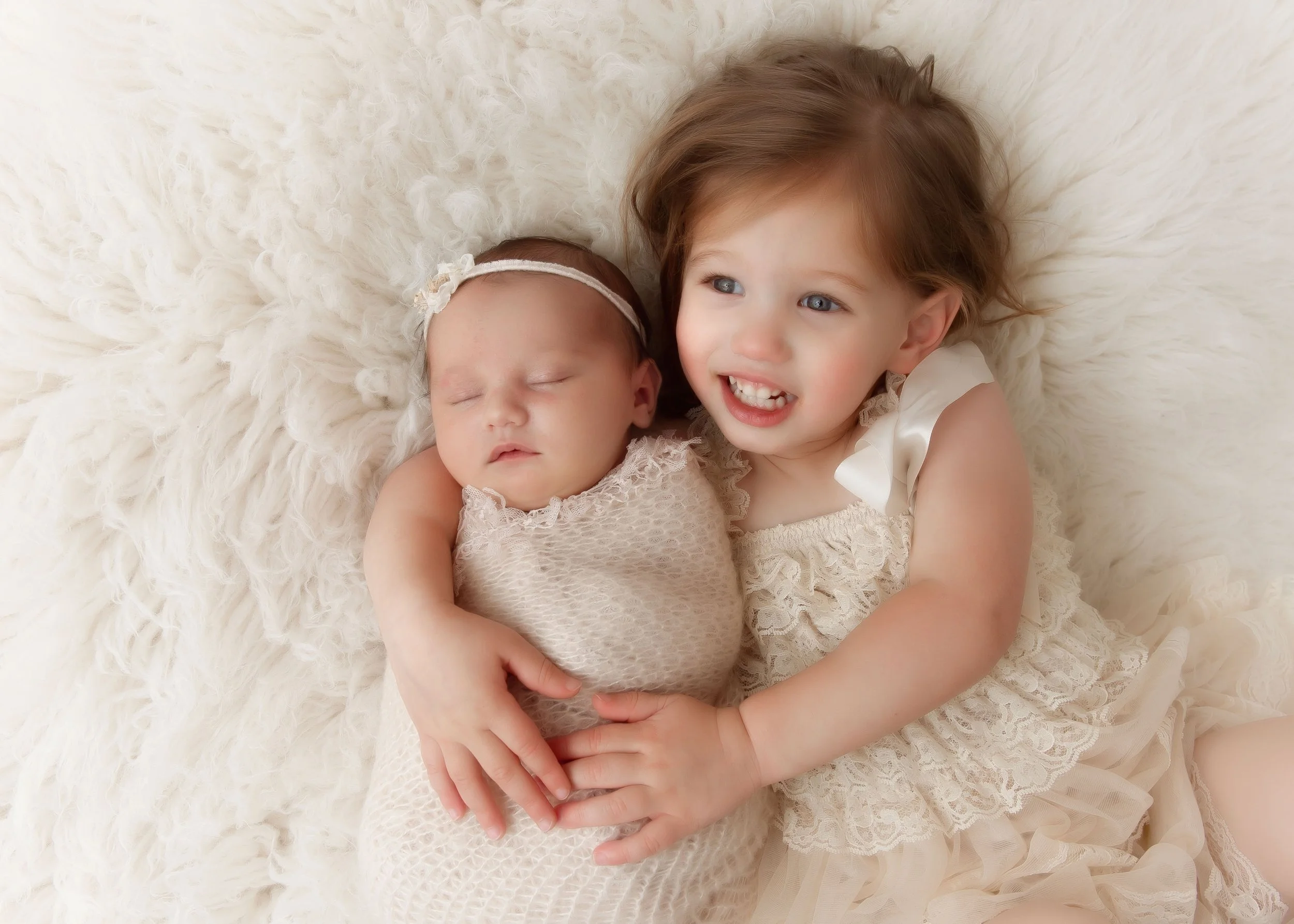 A young girl cuddling a sleeping newborn baby on a fluffy cream-colored blanket.