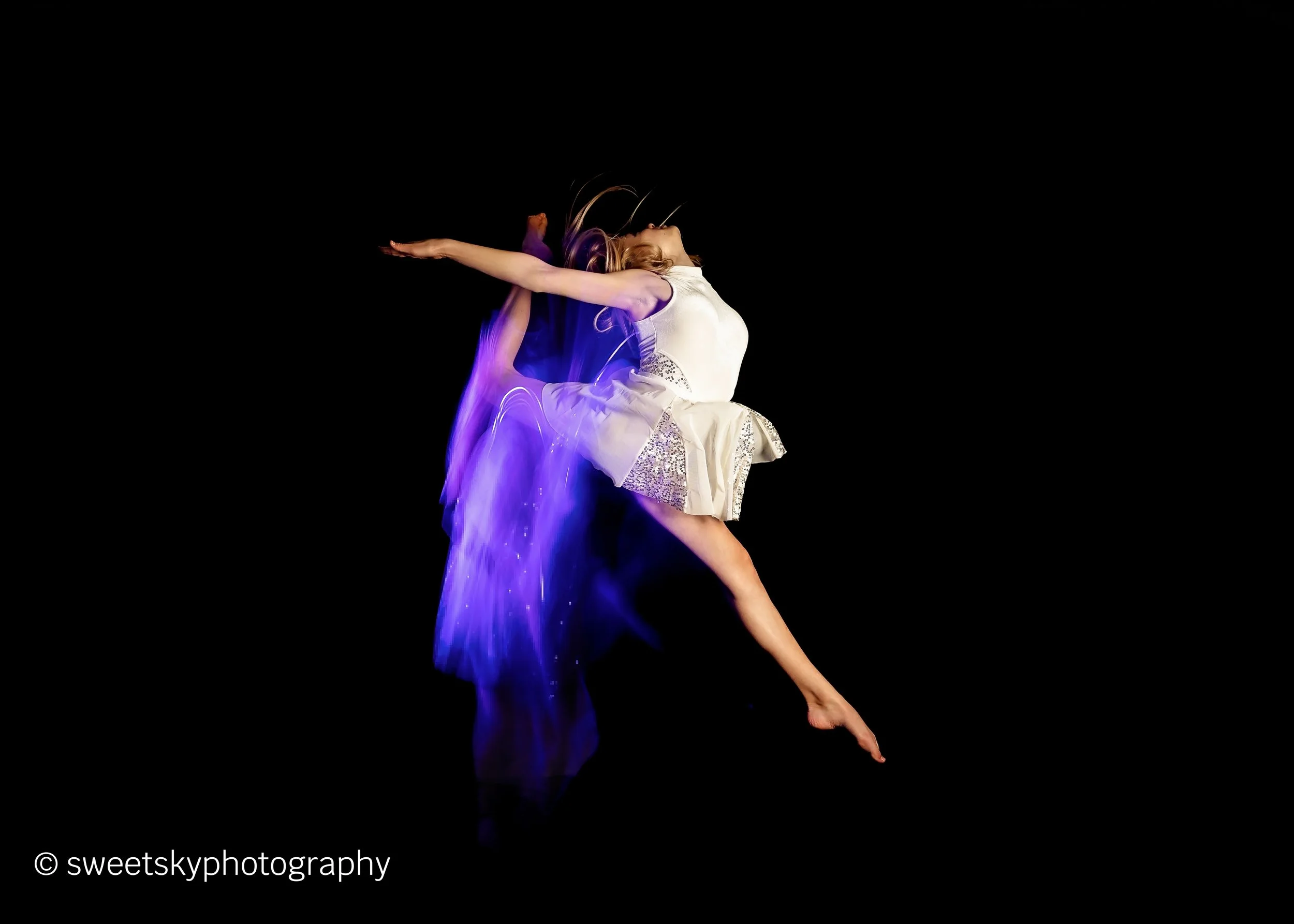 A female dancer in a white dress performing a leap against a black background, with purple light streaks following her motion.