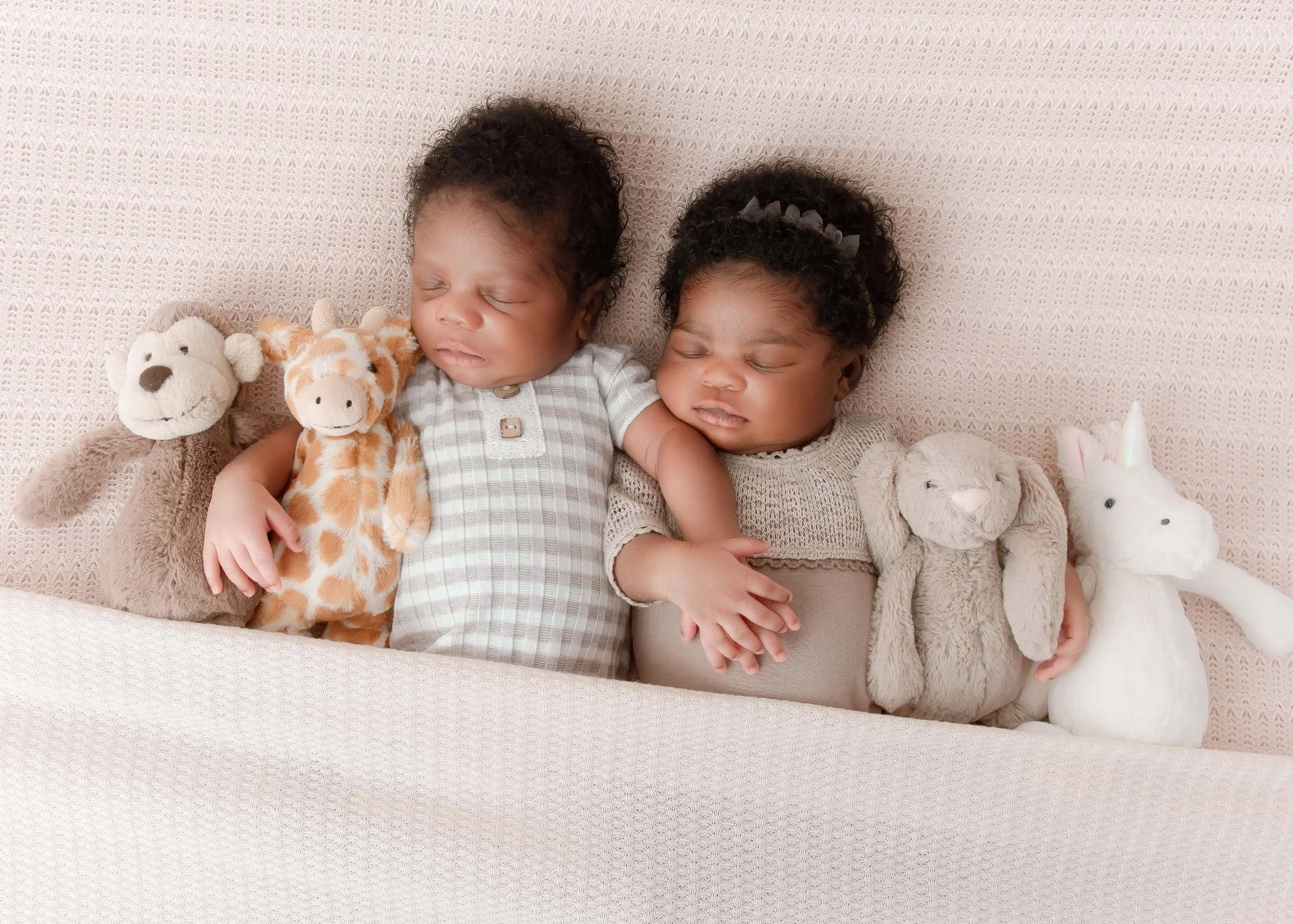 Two sleeping babies lying on a soft pink surface, each holding a plush toy, with additional stuffed animals around them.