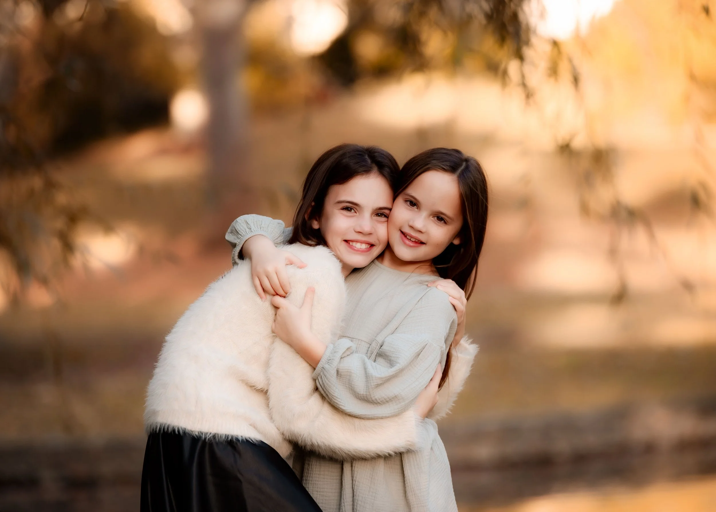Two young girls hugging and smiling outdoors during fall, with blurred trees and a body of water in the background.
