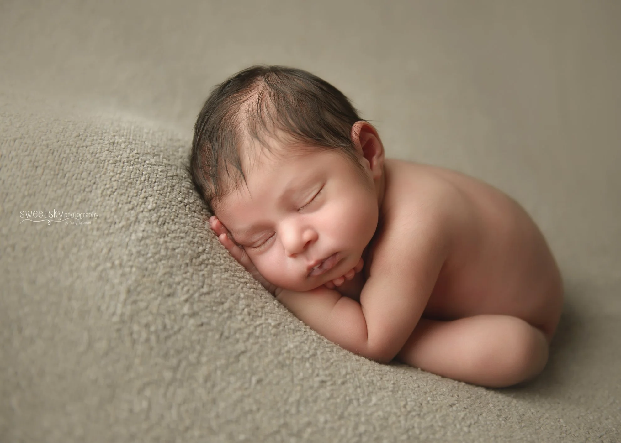 A sleeping newborn baby lying on a textured beige surface with their head resting on their hand, wearing no clothes.