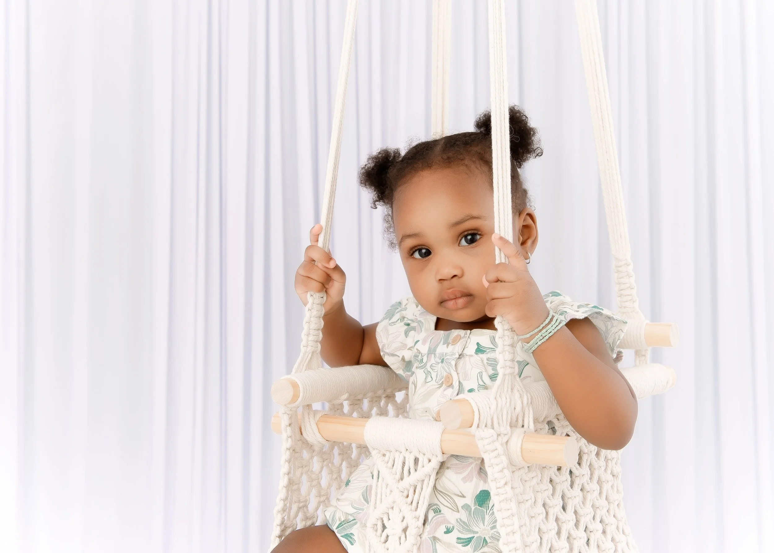 A young girl with curly hair in pigtails sitting in a white macrame swing with a neutral background.