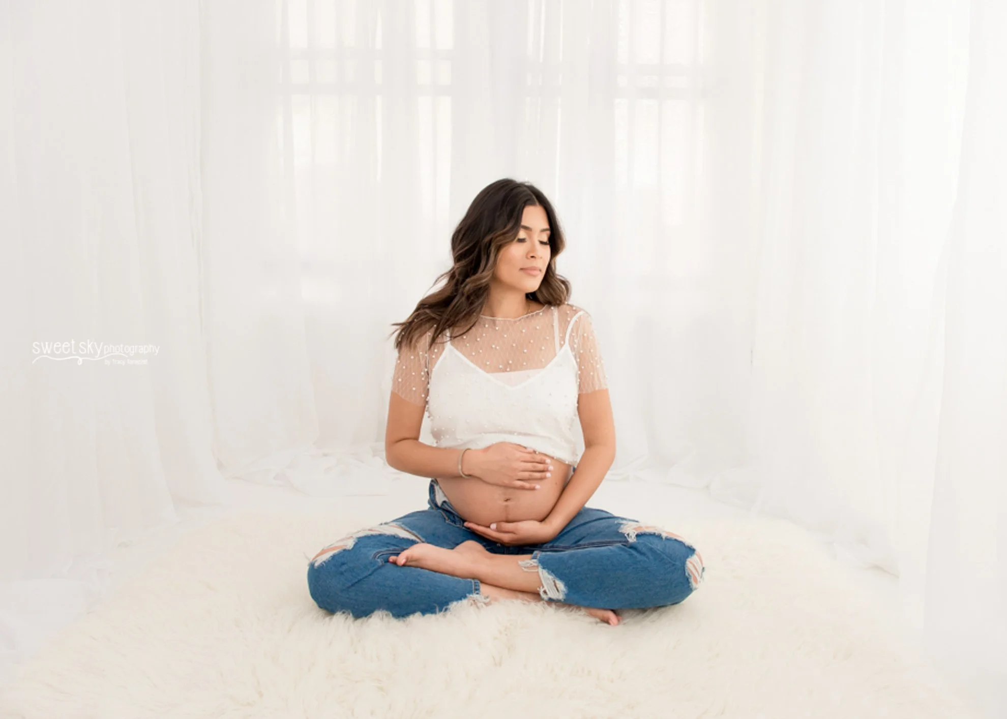 Pregnant woman sitting cross-legged on a white rug, holding her belly with both hands, wearing a sheer polka dot top and ripped jeans, in a bright room with white curtains.