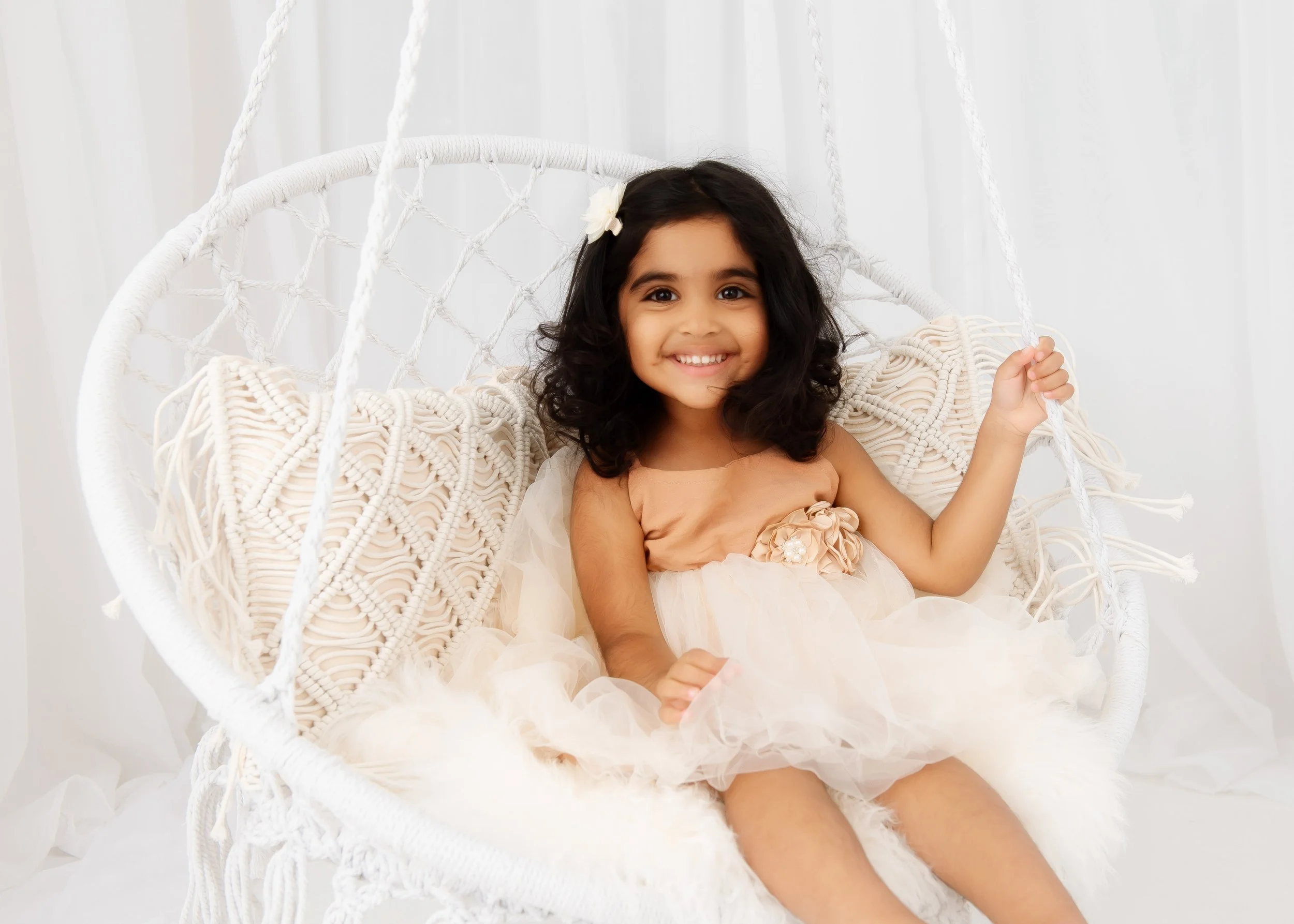 A young girl with dark curly hair sitting on a white woven swing chair, smiling at the camera, wearing a peach-colored dress with floral embellishments, in a bright room with white curtains.