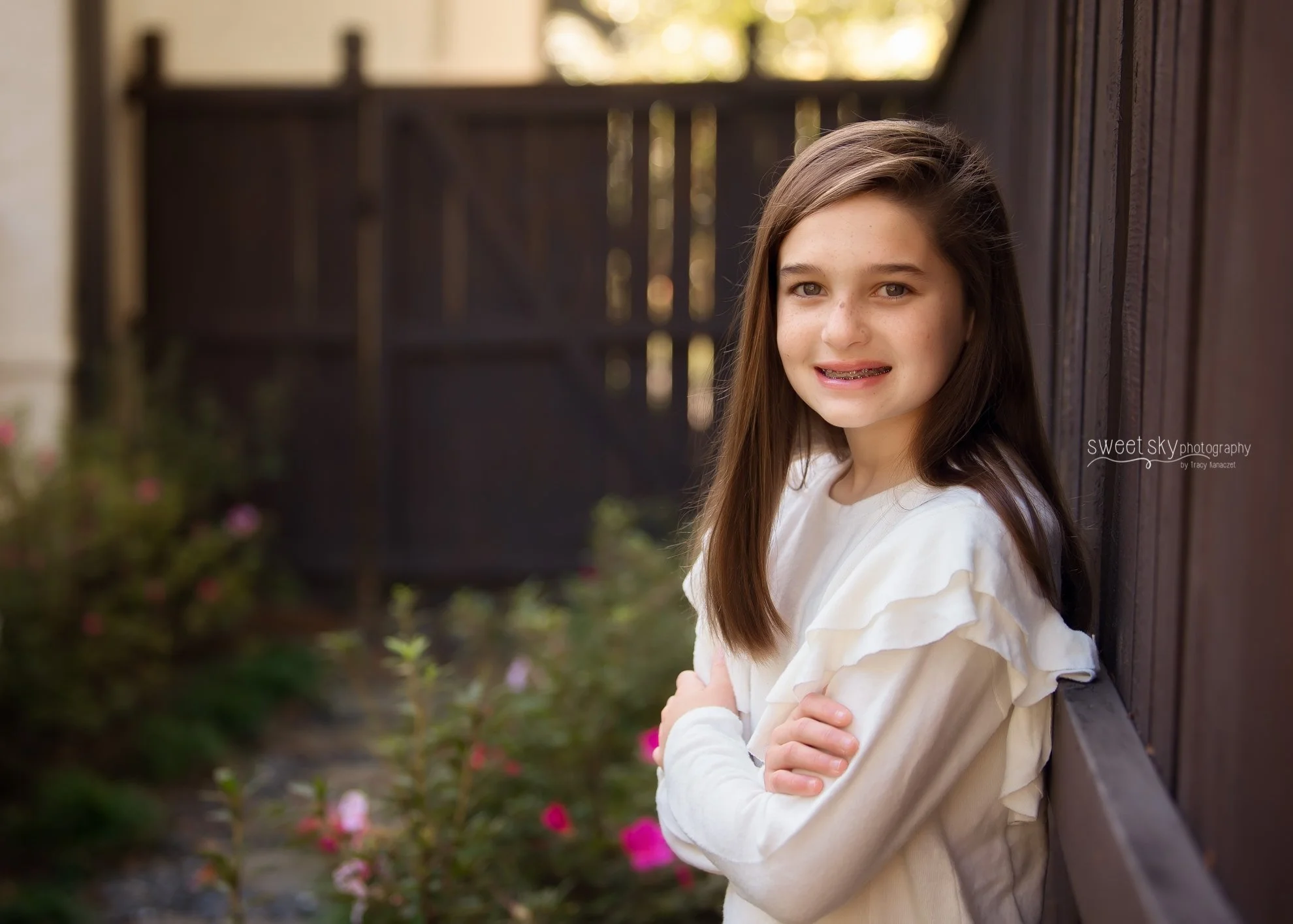 Young girl with long brown hair and braces, wearing a white ruffled sweater, leaning against a dark wooden fence with arms crossed, smiling at the camera, with flowering plants in the background.