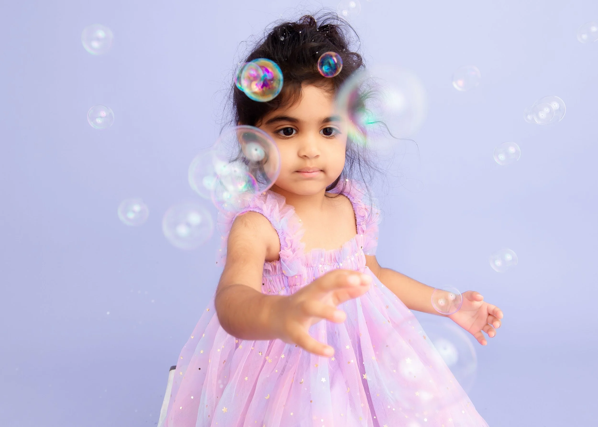 A young girl in a pink princess dress reaching out towards floating soap bubbles against a light purple background.