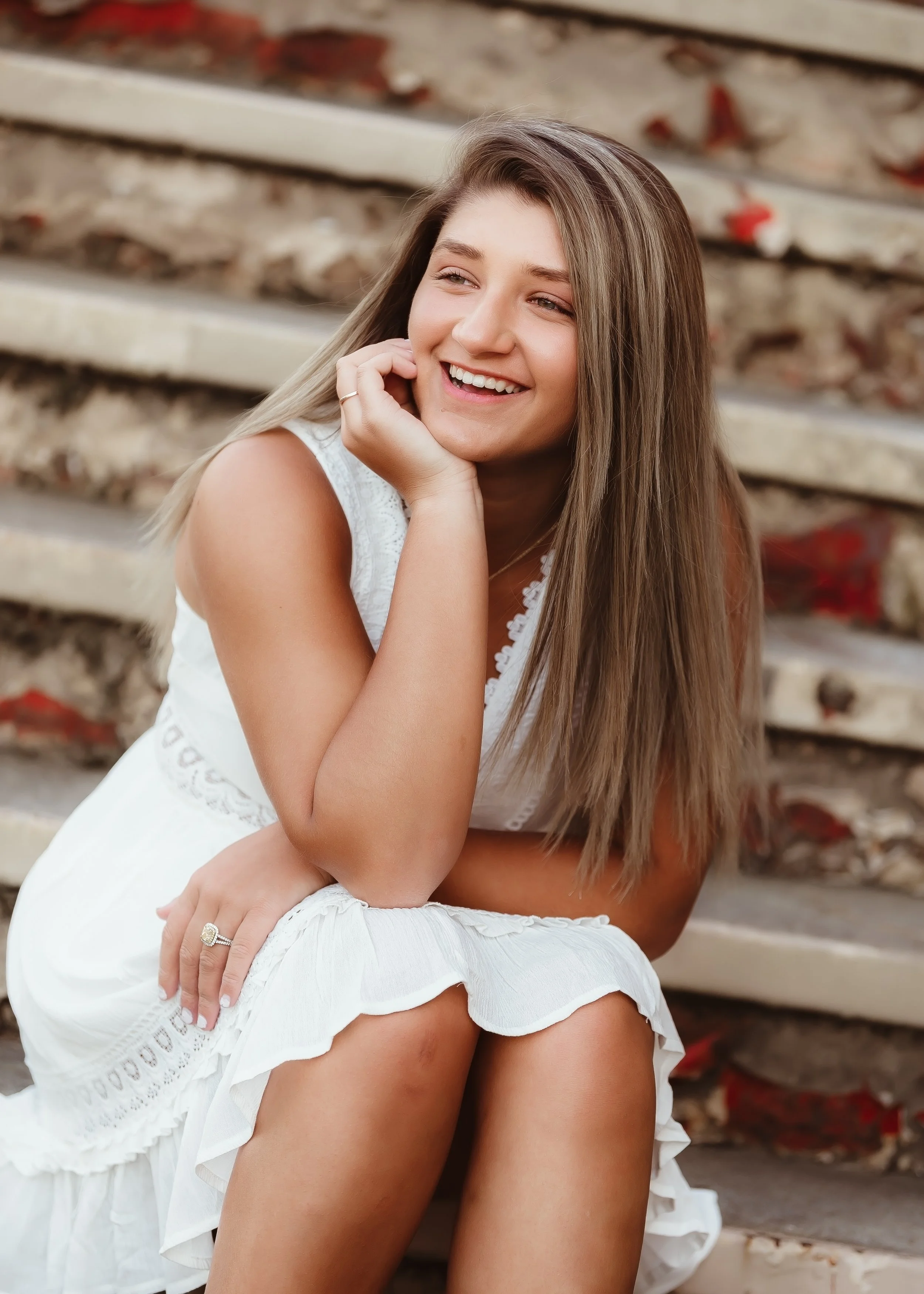 A young woman with long, light brown hair sitting on outdoor stone stairs, smiling and wearing a white sleeveless dress.