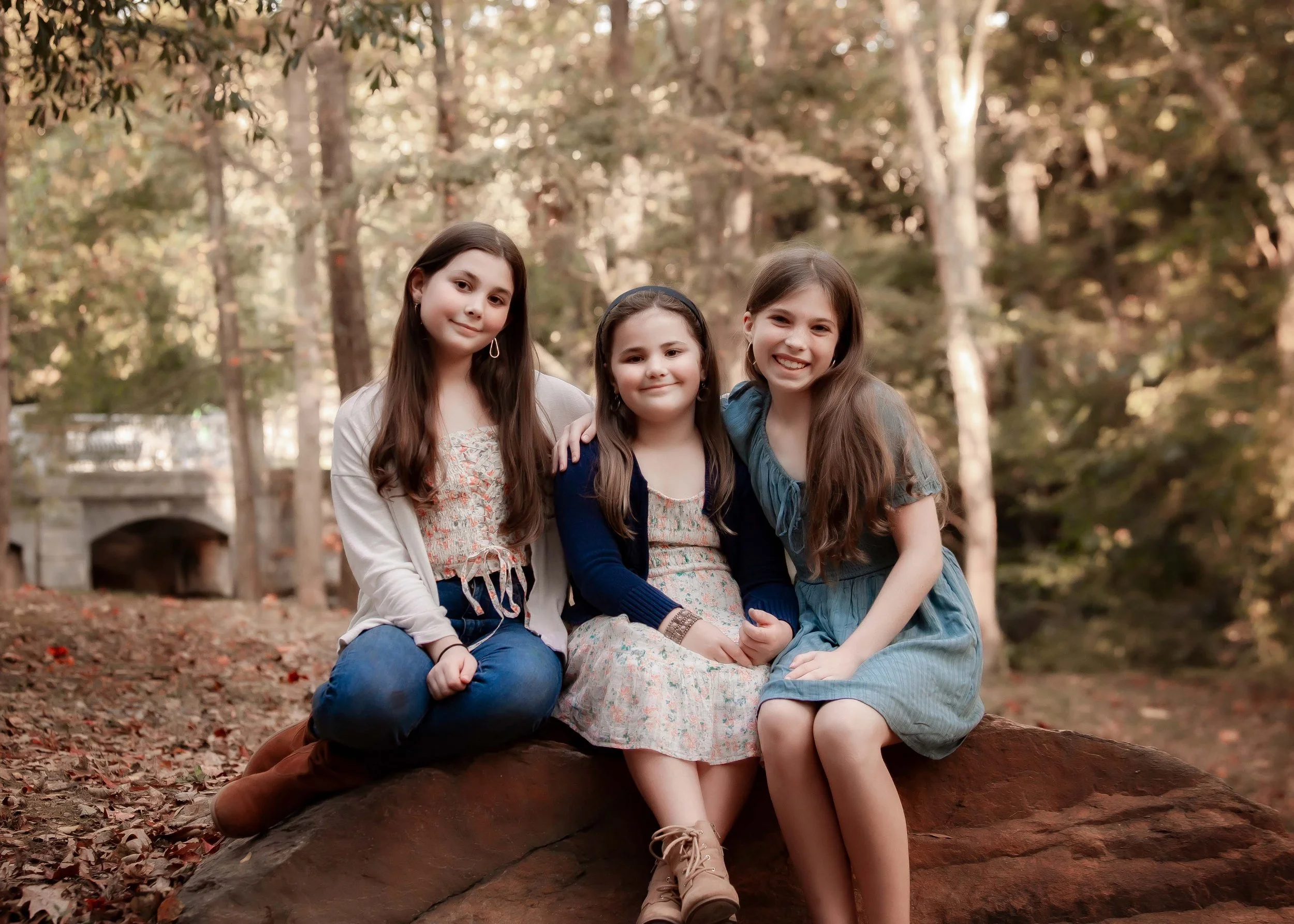 Three young girls sitting on a large rock in a wooded area, smiling at the camera.