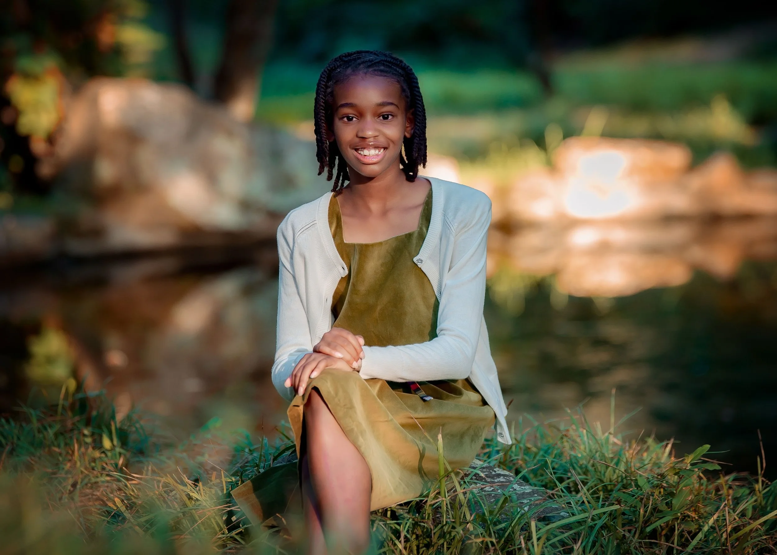 A young girl sitting outdoors on a log near a body of water, smiling at the camera, with blurred trees and rocks in the background.