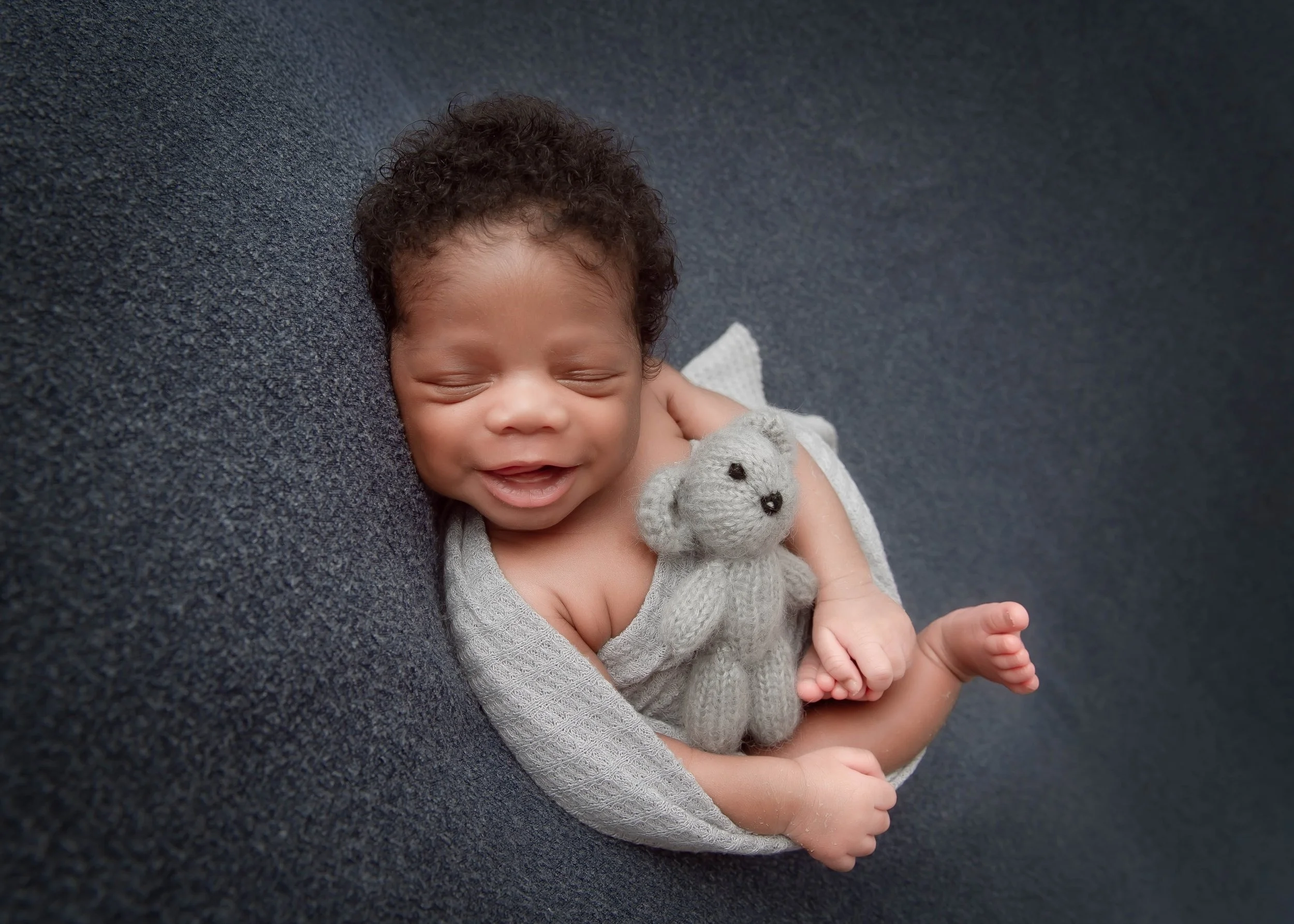 Baby lying on a dark grey surface, smiling with eyes closed, holding a small knitted grey teddy bear.
