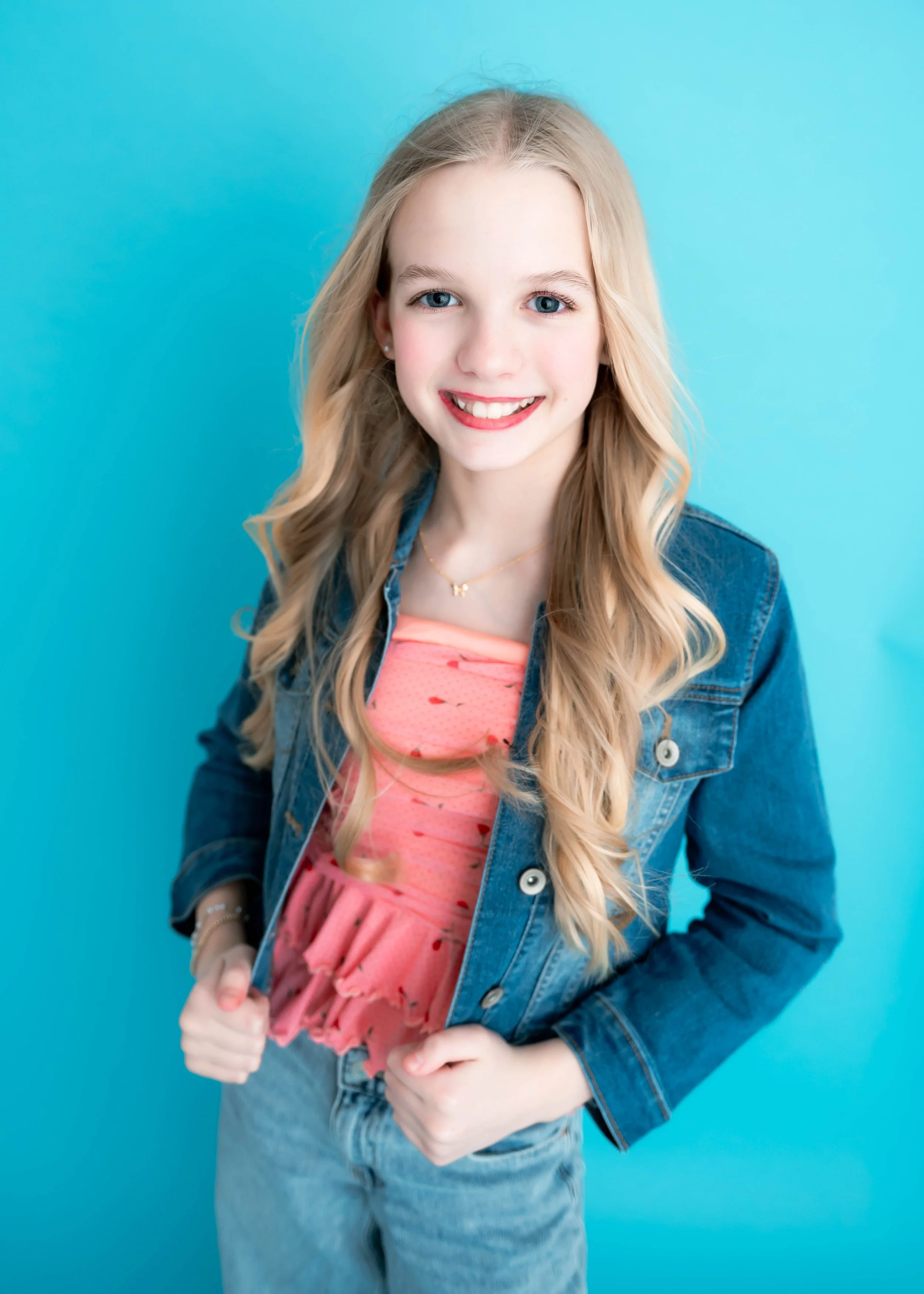 Young girl with long blonde hair, smiling, wearing a denim jacket over a pink dress, standing against a blue background.