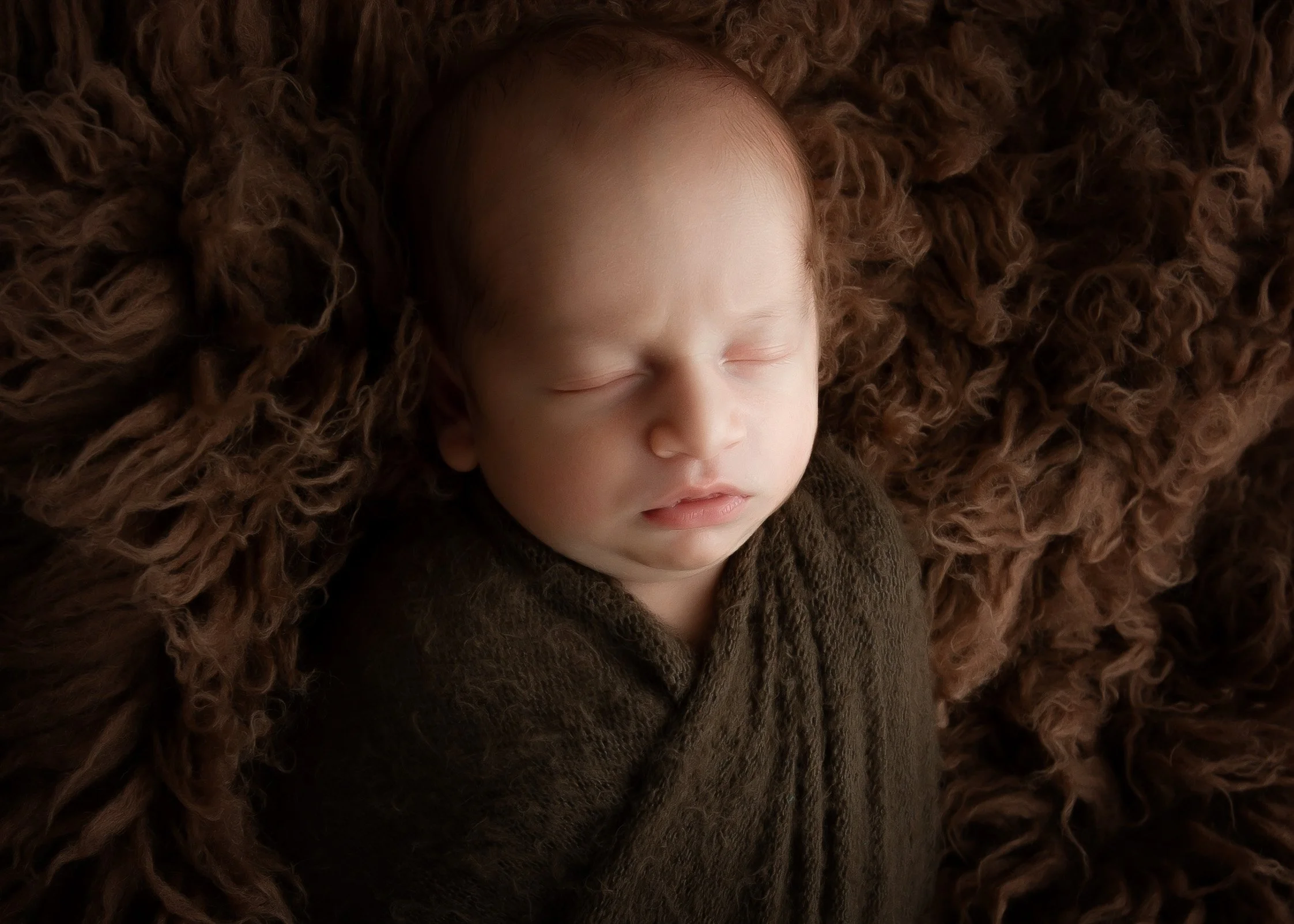 A sleeping baby with closed eyes lying on a brown, fluffy blanket, wearing a dark brown outfit.
