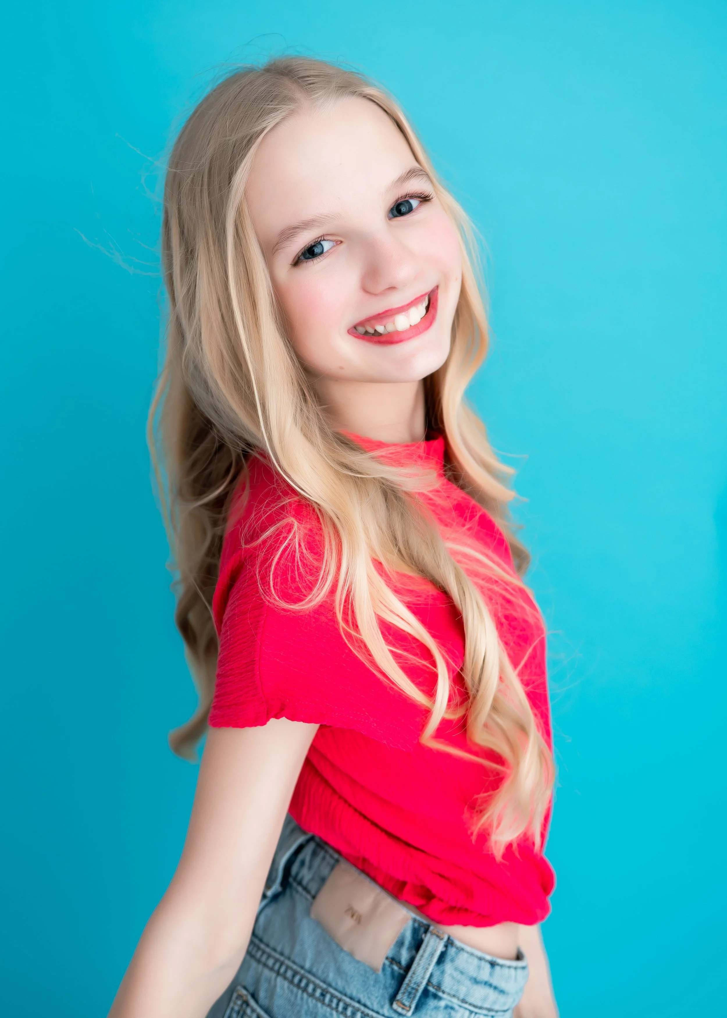 A young girl with long blonde hair styled in loose waves, wearing a bright pink t-shirt and denim shorts, smiling against a blue background.