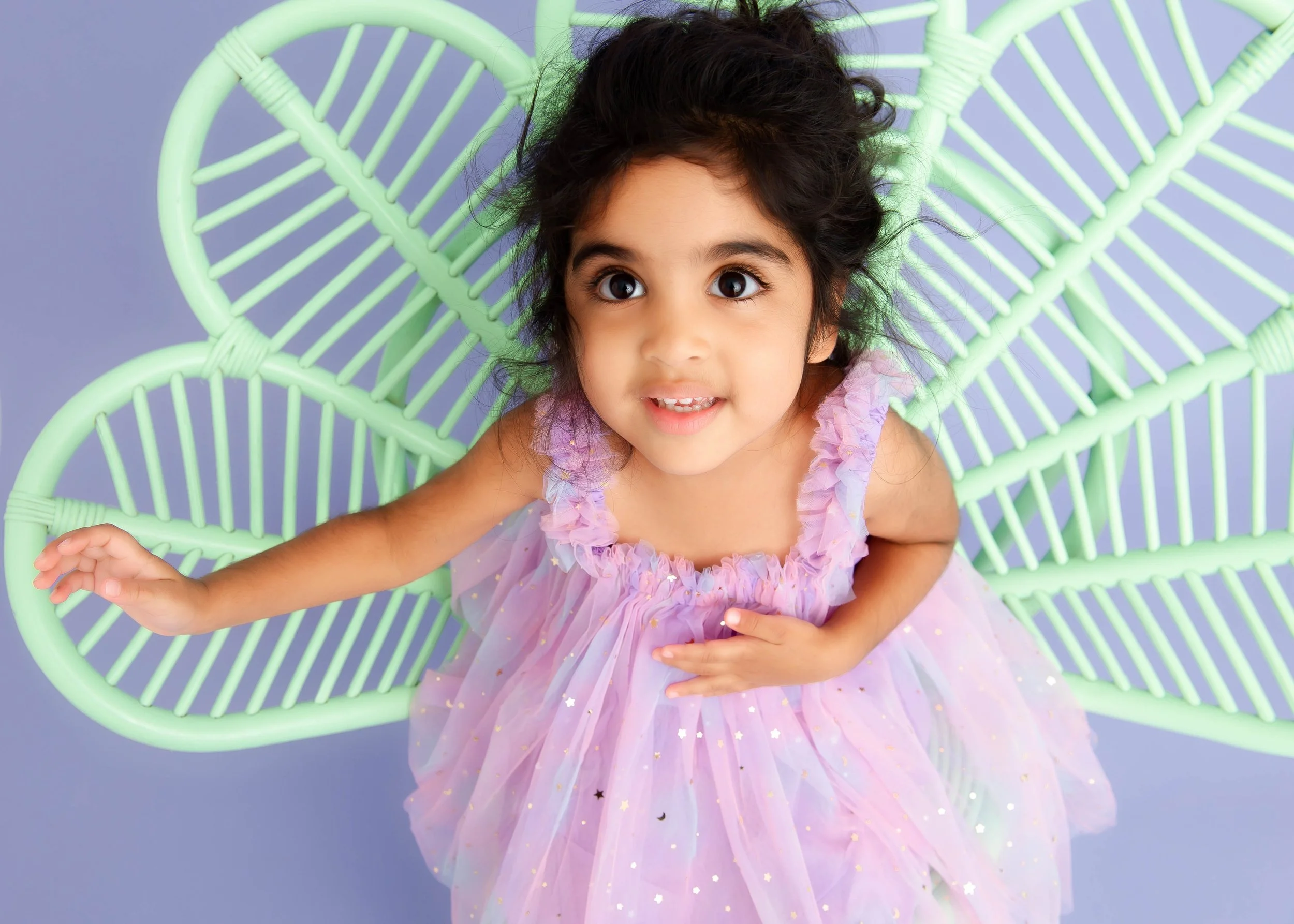 A young girl with dark hair and big eyes, wearing a pastel purple dress with ruffled sleeves and star-shaped sequins, sitting on a light green wire chair, looking up with a surprised expression.