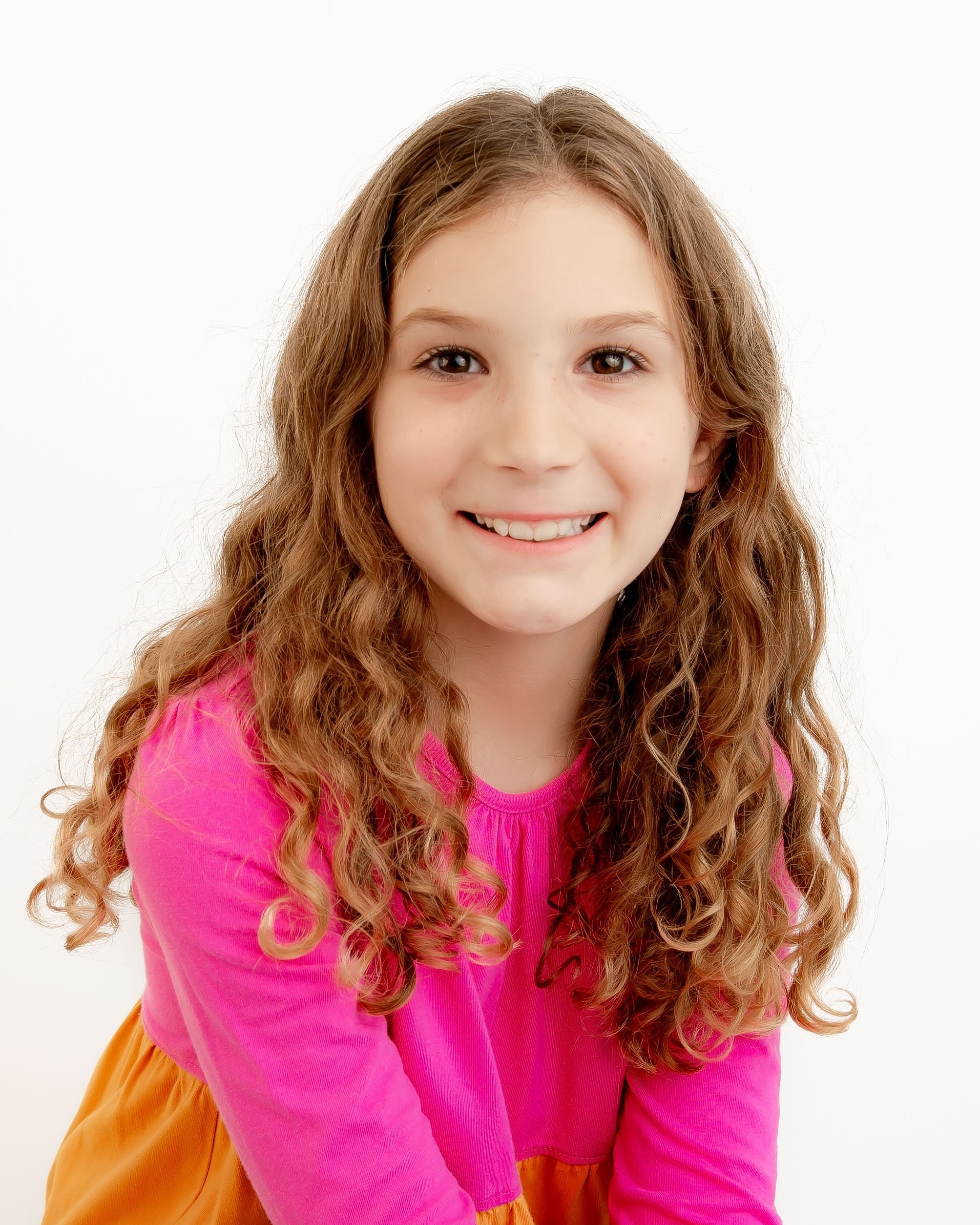 Young girl with long curly hair smiling, wearing a bright pink top and orange skirt, against a white background.