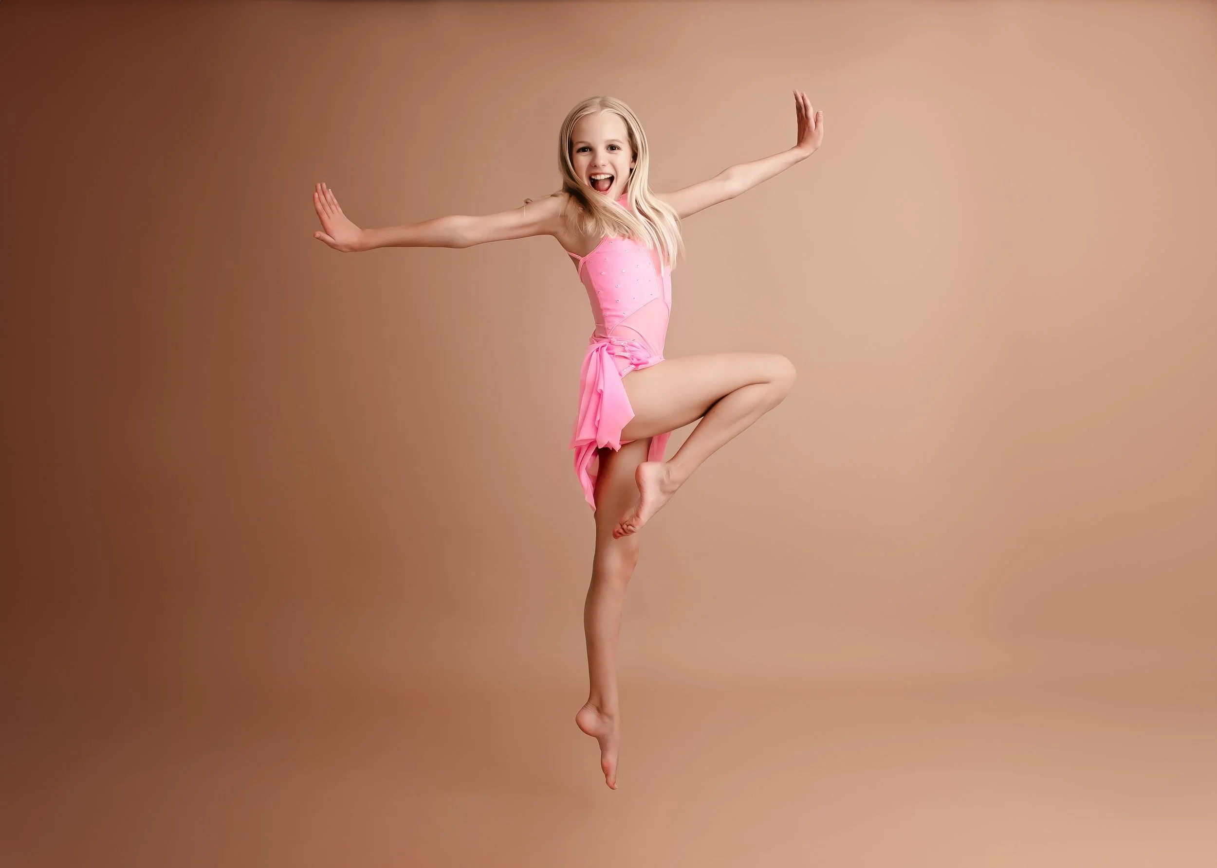 A young girl in a pink ballet dress jumping with arms outstretched and a big smile, against a neutral background.