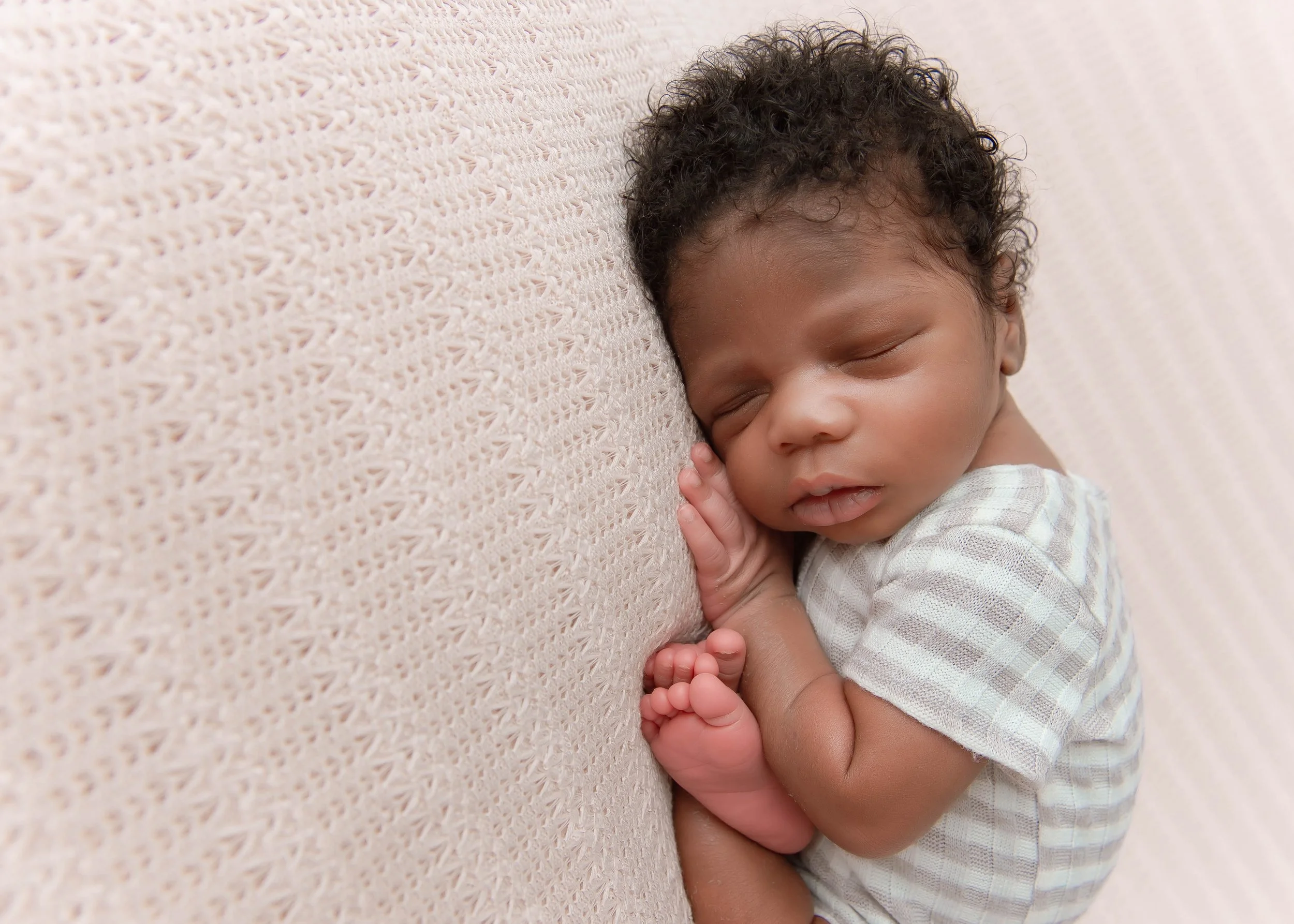A sleeping baby with curly hair, resting against a textured beige pillow or blanket, wearing a light-colored striped shirt.