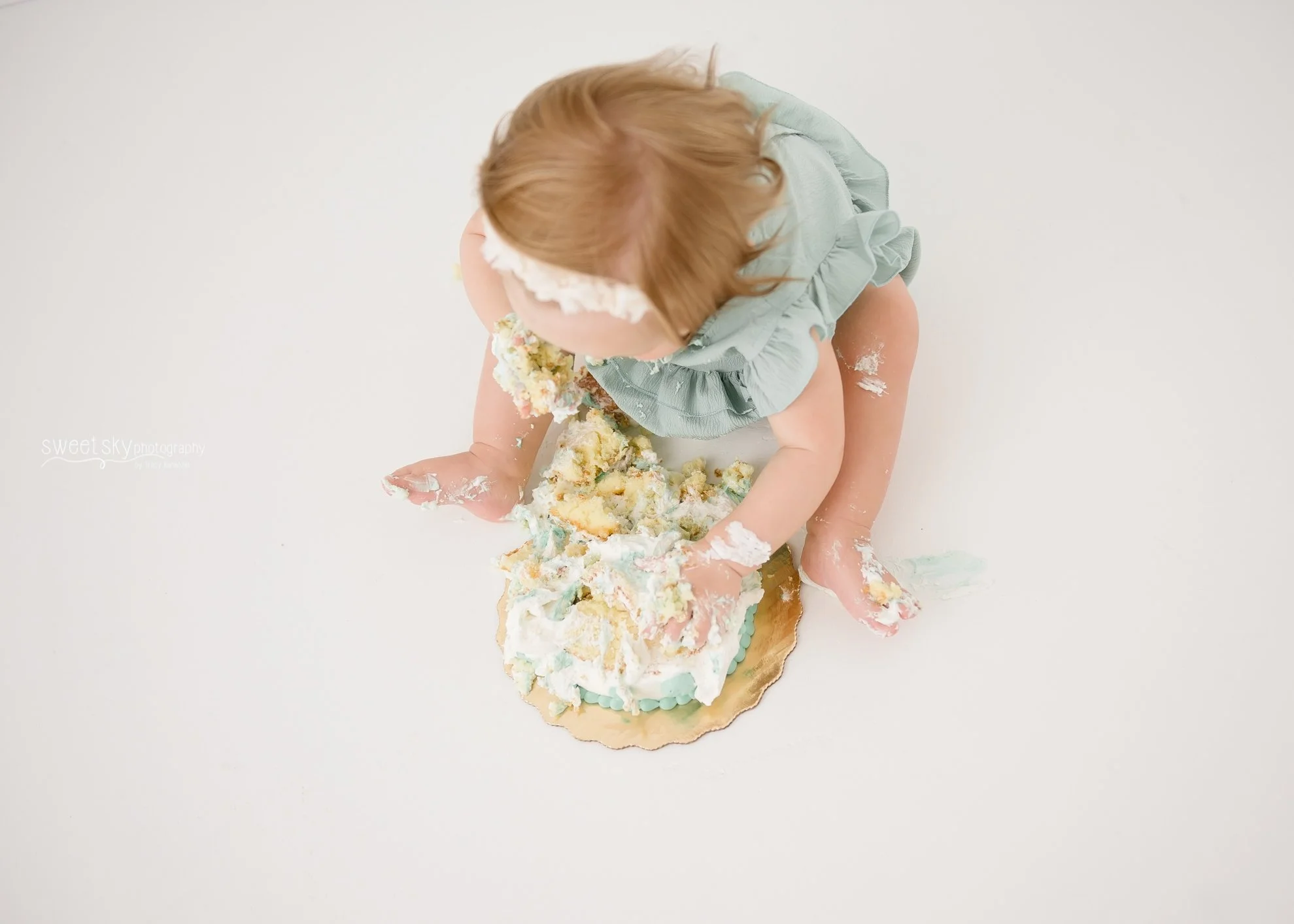A baby sitting on a white floor covered in cake and icing, wearing a light blue dress and a floral headband, with a partially eaten birthday cake in front of her.