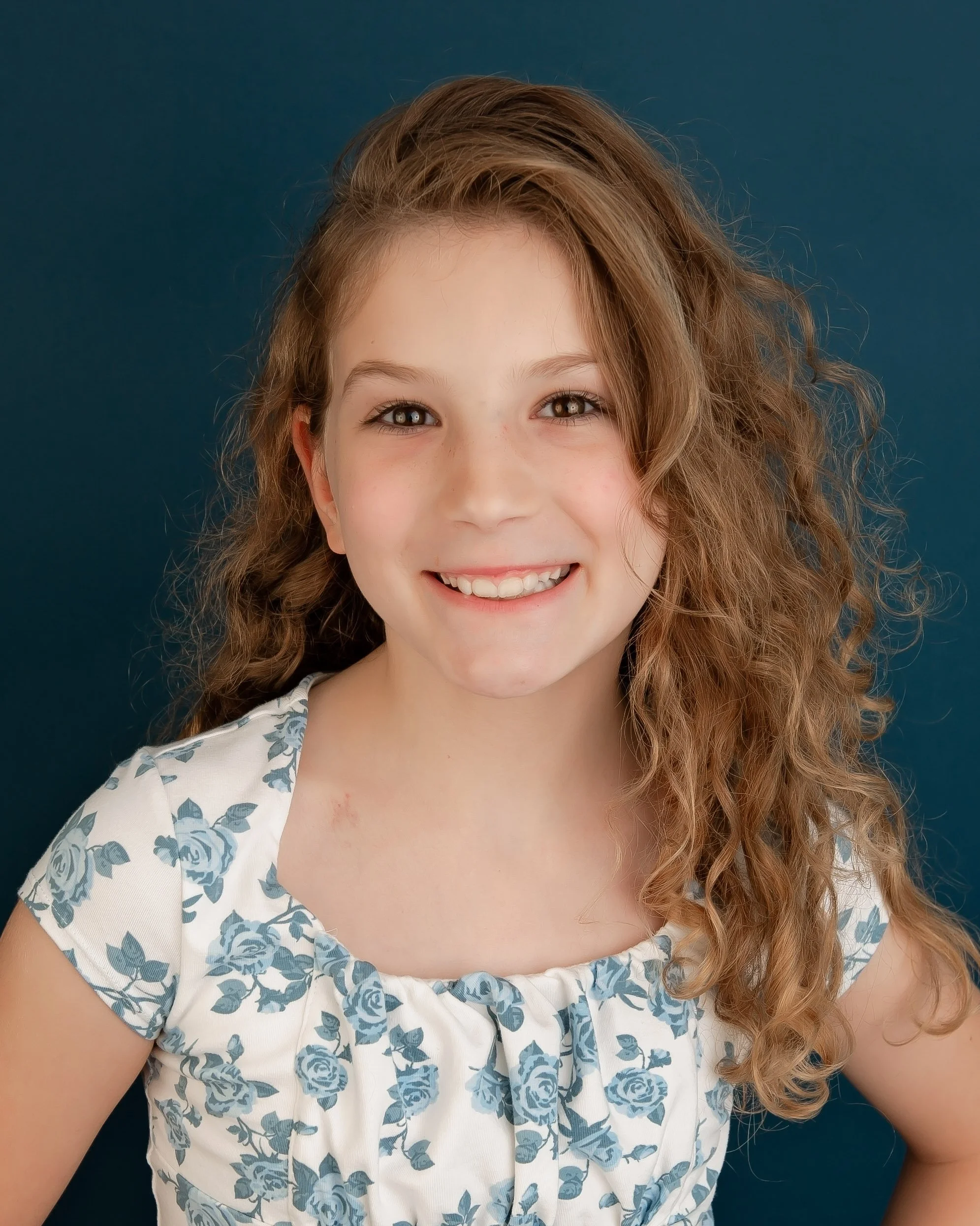 Smiling young girl with long, curly, light brown hair, wearing a white dress with a blue floral pattern, posed against a dark blue background.